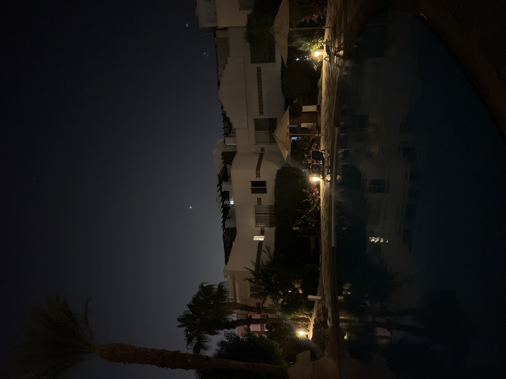 A nighttime exterior shot captures a resort swimming pool area in Sharm El Sheikh, Egypt. A large, dark swimming pool dominates the foreground, reflecting the ambient light from the surroundings. In the mid-ground, a paved poolside area features several empty lounge chairs and two open umbrellas. Landscape lighting illuminates the area, casting a warm glow on tropical foliage and the white exterior of resort buildings situated in the background. Palm trees are visible on the left and in the mid-ground, their silhouettes stark against the dark night sky, which is faintly dotted with a few stars. The resort buildings appear to be two or three stories high with visible windows and architectural details. The scene is serene and unoccupied, conveying a sense of peaceful relaxation.