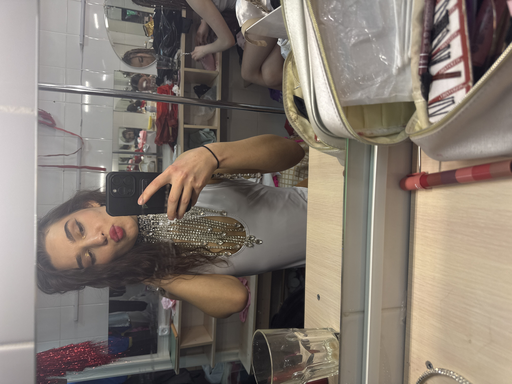 The image shows a person with brown hair and red lipstick taking a selfie in a mirror. The person is wearing a white top with a silver, jewel-encrusted neckline. The background appears to be a dressing room or backstage area with a mirror reflecting the scene, a makeup bag, and various items on shelves. There is a glass of water on a counter near the mirror.
