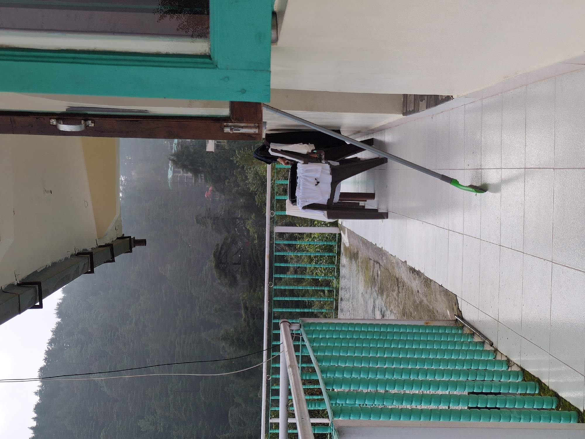 This is a vertical, medium-shot, eye-level outdoor photograph taken from a balcony overlooking a misty, forested hillside in Bhagsu, India. The balcony floor is tiled with large, off-white squares, wet from recent rain. A turquoise metal railing runs along the left edge of the balcony, with a white handrail above it. Beyond the railing, the misty green slopes of a mountain are covered in dense coniferous trees.
On the balcony, two brown plastic chairs are placed near the wall. One chair is partially obscured by a dark blue garment, possibly a piece of clothing, draped over its back. A white shirt or cloth is spread across the seat of the same chair. Leaning against the wall is a mop with a green base and a long grey handle. The wall of the building is painted a light cream color. To the right, a portion of a window frame, painted turquoise, is visible. The window itself reflects the overcast sky and the misty environment. The scene suggests a quiet, damp day in the mountainous region.