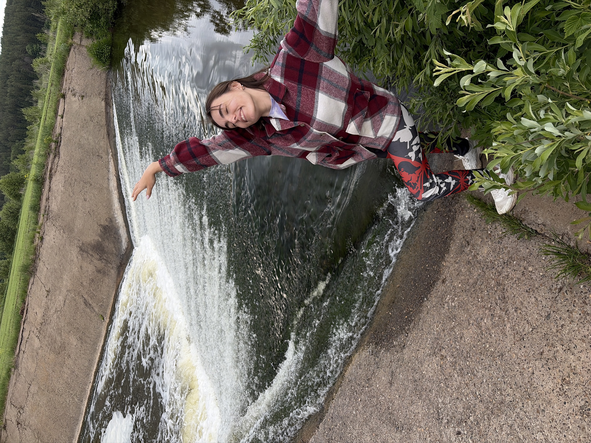 The photograph captures a woman standing near a waterfall in what appears to be a rural or park-like setting. She has light brown hair and is wearing a red and white plaid shirt, black, red, and white leggings, and white sneakers. The woman is smiling and has her arms outstretched as if posing for the photo.
Behind her, a concrete dam or structure directs the flow of water over the edge to create the waterfall. Green grass is visible along the side of the dam. To her right, there are trees and vegetation, some of which she seems to be standing close to.
The image is rotated counter-clockwise. The location is Nizhnii Vishur, Russia.