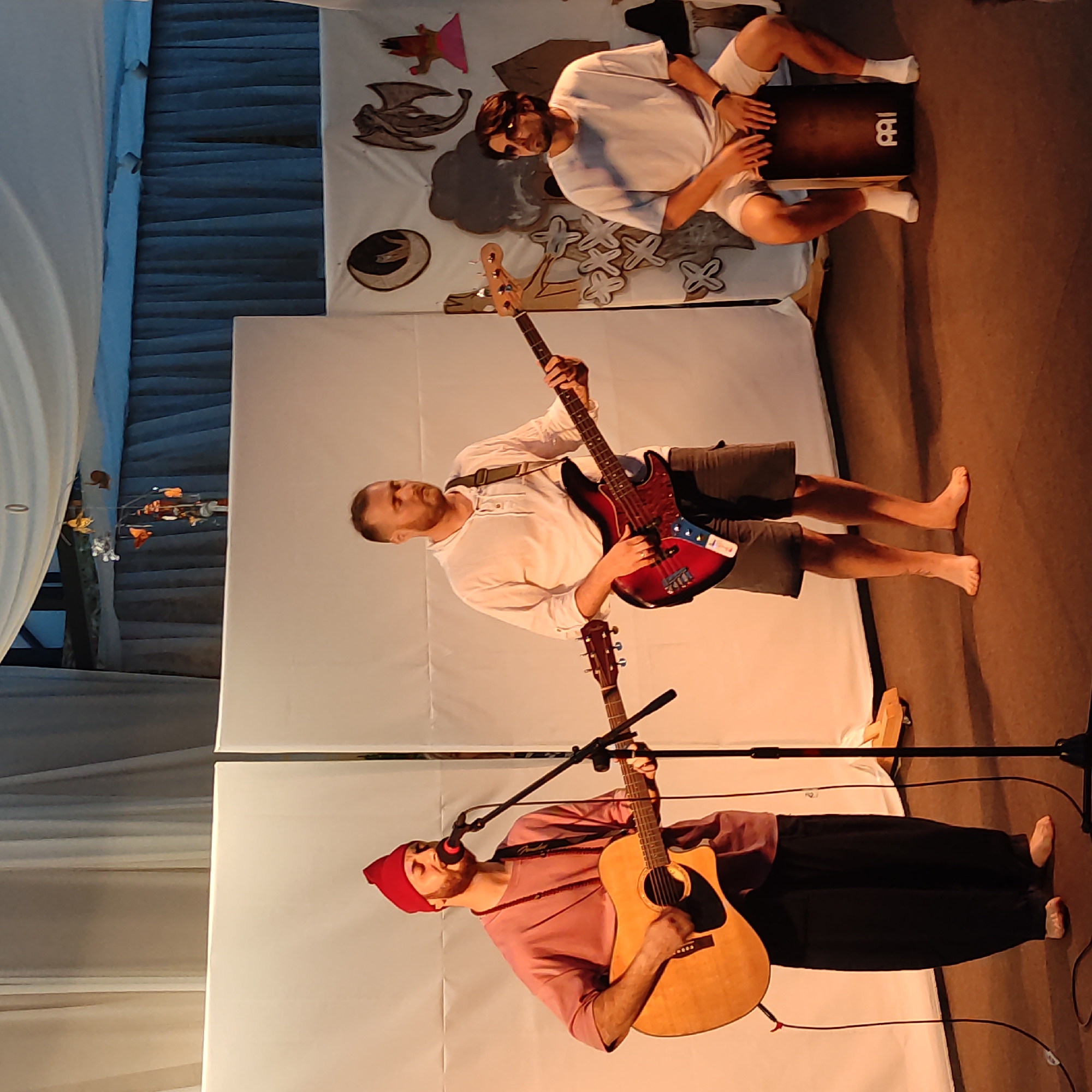 A group of three men are performing music on a stage in Zhirnosovo, Russia. The man on the left is wearing a red beanie and a pink shirt and is playing an acoustic guitar and singing into a microphone. The man in the middle is wearing a white long-sleeved shirt and is playing a red and black bass guitar. The man on the right is wearing a white t-shirt and shorts and is sitting on a cajón drum, playing it with his hands. The stage has a white backdrop with some decorative elements, including a moon graphic, a dragon graphic, and other cutouts. The lighting appears to be warm and theatrical. The floor is covered in a dark brown carpet.