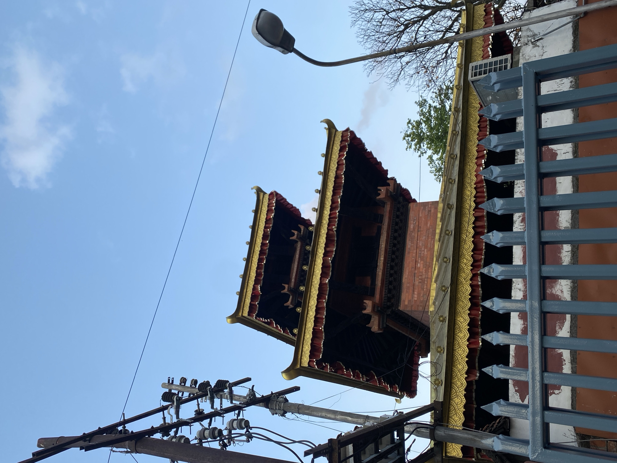 The image captures a tiered, pagoda-style building with golden eaves and red and gold fabric draping. The structure appears to be made of brick. In the foreground, a grey metal fence with pointed finials runs horizontally across the lower portion of the frame. To the left, a tall, weathered utility pole with electrical wires and insulators stands prominently. A streetlamp with a curved pole is visible on the right side of the image, with a bare-branched tree and some green foliage behind it. The sky above is a pale blue with scattered clouds. The architectural style suggests the location is likely within Kathmandu Metropolitan City, Nepal, possibly a temple or cultural heritage site. There are no people visible in the image.