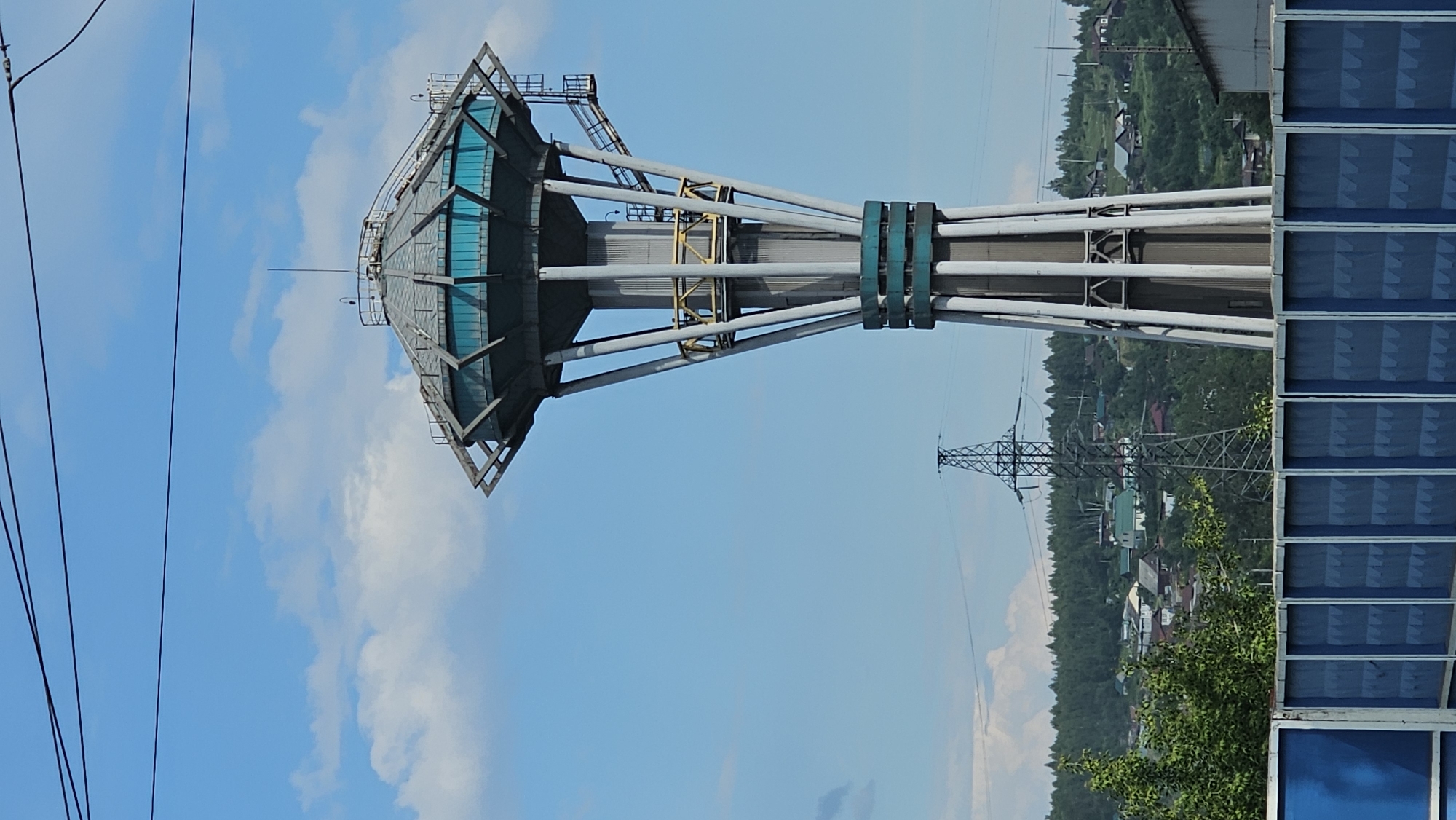 This media file captures an outdoor scene in Revda, Russia, dominated by an industrial structure that resembles a water tower or a cooling tower, with a distinctive hexagonal top featuring blue panels. The tower is supported by multiple large, angled pipes, some of which are banded with green. The surrounding landscape is lush with green trees, and a small village with houses and red roofs is visible in the distance. A tall, lattice-style electrical pylon stands prominently in the mid-ground, indicating the presence of industrial infrastructure. A blue, glass-paneled building facade is visible on the right side of the frame, adding a modern architectural element. The sky is a clear blue, dotted with white clouds, suggesting a pleasant daytime with good weather. Several power lines are also visible, stretching across the left side of the image. There is no visible text in the image. The overall impression is one of a functional, industrial area set against a natural and semi-urban backdrop.