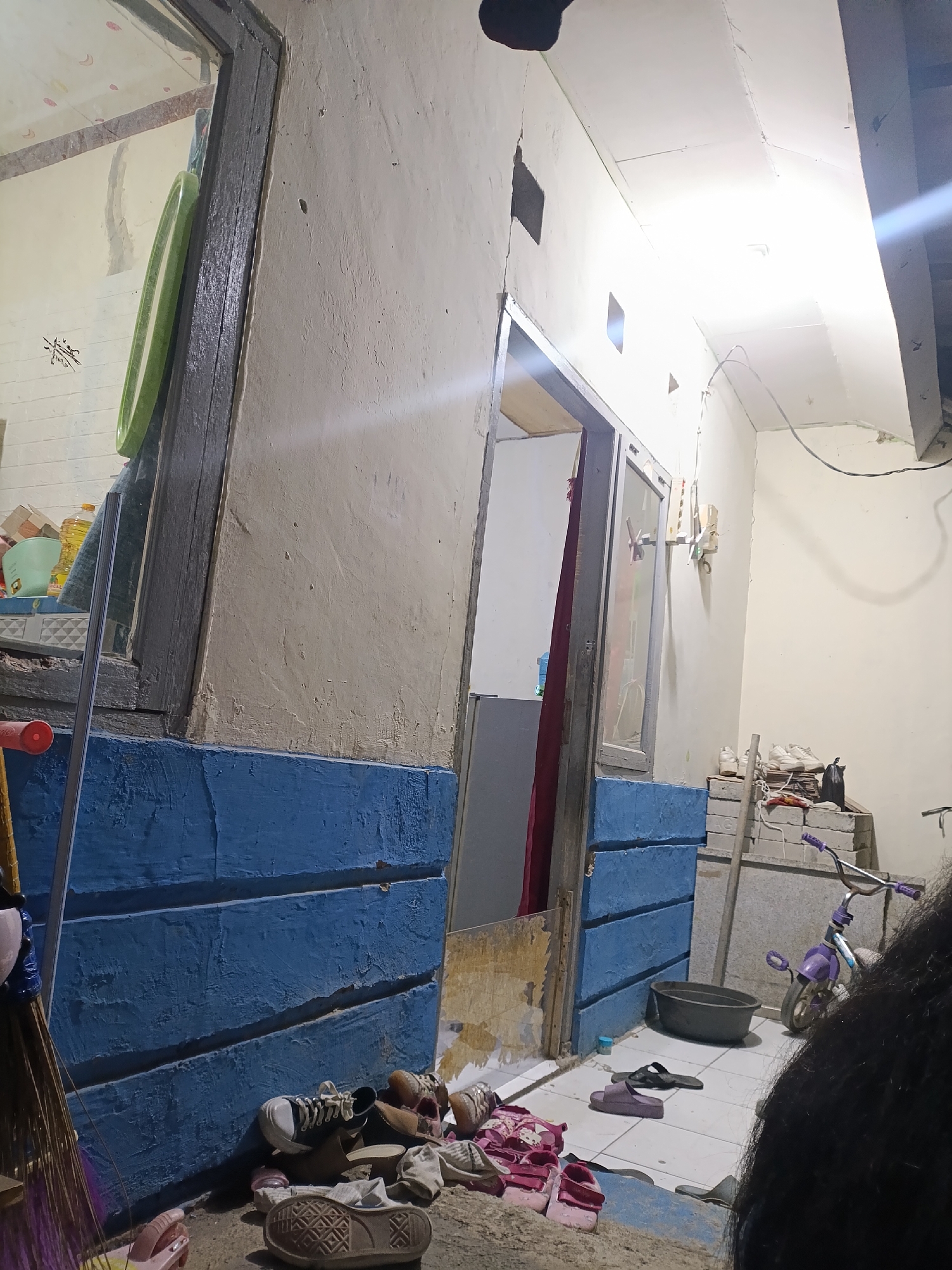 The image depicts an outdoor scene, likely a backyard or alleyway, in Cariu, Indonesia. The ground is tiled, and there is a blue painted wall with visible horizontal planks. A doorway is visible, leading into a dimly lit interior. Various objects are scattered around, including shoes, sandals, a broom, a child's bicycle, and a dark grey basin. The wall of the structure appears to be textured and aged, with some peeling paint. The lighting suggests it may be dusk or that artificial light sources are present but not fully illuminating the area. The presence of domestic items suggests a residential setting.
