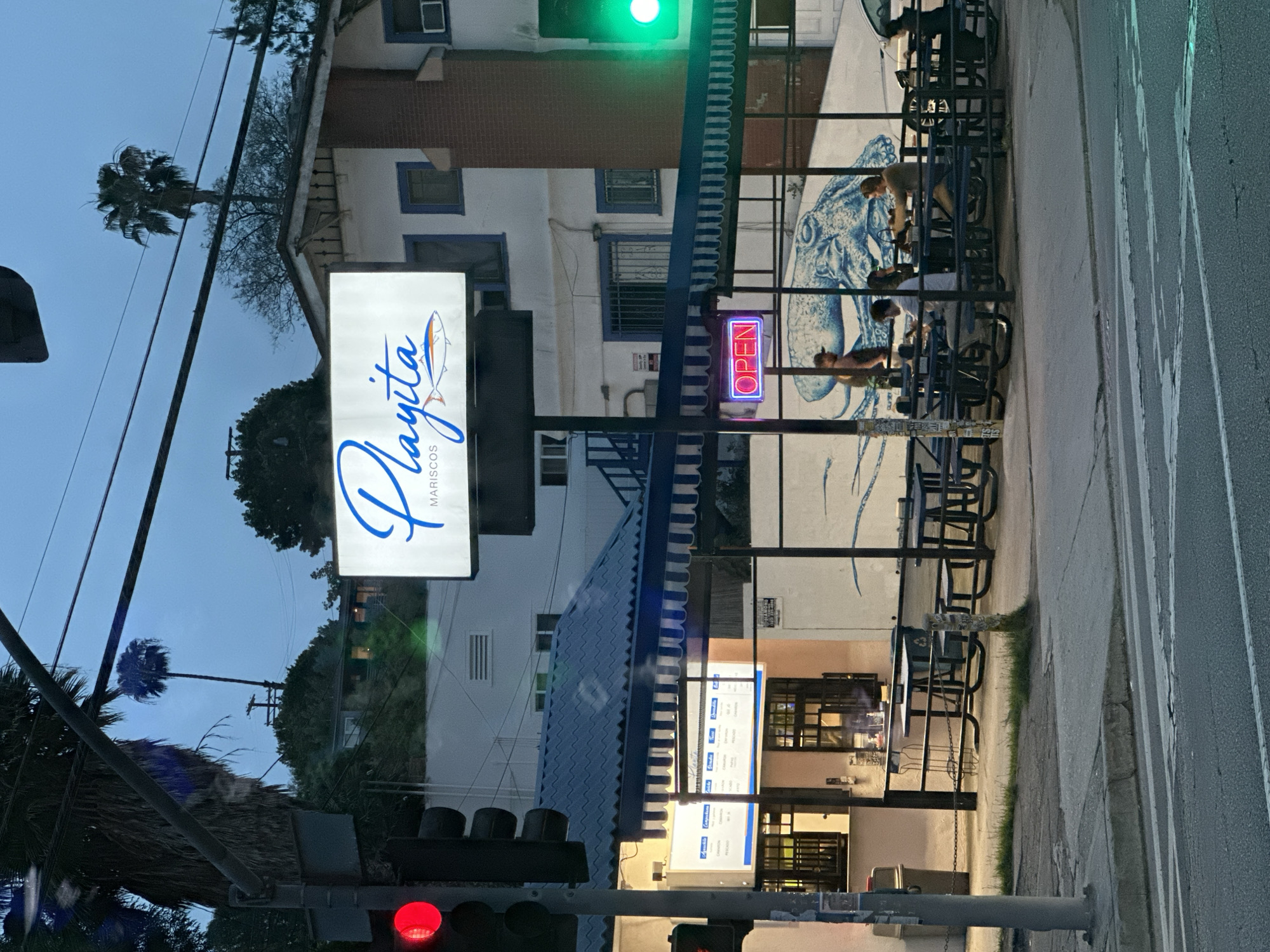 The image depicts the exterior of "Playita Mariscos," a seafood restaurant. The restaurant has outdoor seating with a mural of an octopus on the wall behind the tables. A sign with the restaurant's name in blue script is visible, and an "OPEN" neon sign is lit. There is a building with white and blue accents across the street from the restaurant. The image appears to be taken at dusk. The location is Los Angeles, California. The traffic light is red.