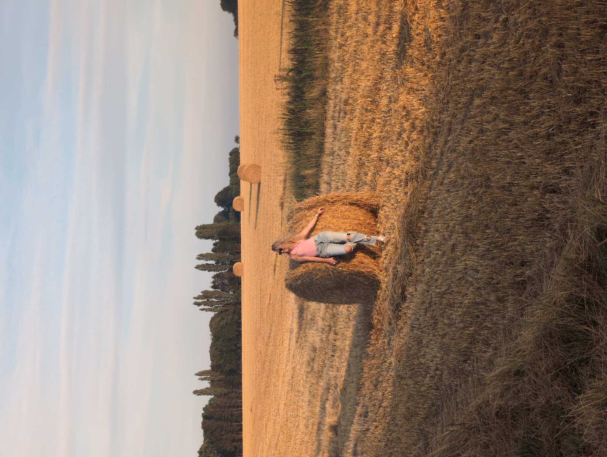 In Alvimare, France, a young Caucasian woman with blonde hair stands leaning against a round bale of hay in a field of dry, cut wheat. She is wearing a pink tank top and ripped blue jeans. Her sunglasses are on her head. The field is filled with rows of hay bales, some closer and more detailed, others further in the background. A line of trees forms the horizon behind the field, under a pale blue sky with wispy clouds. The sun is low, casting long shadows and bathing the scene in a warm, golden light.