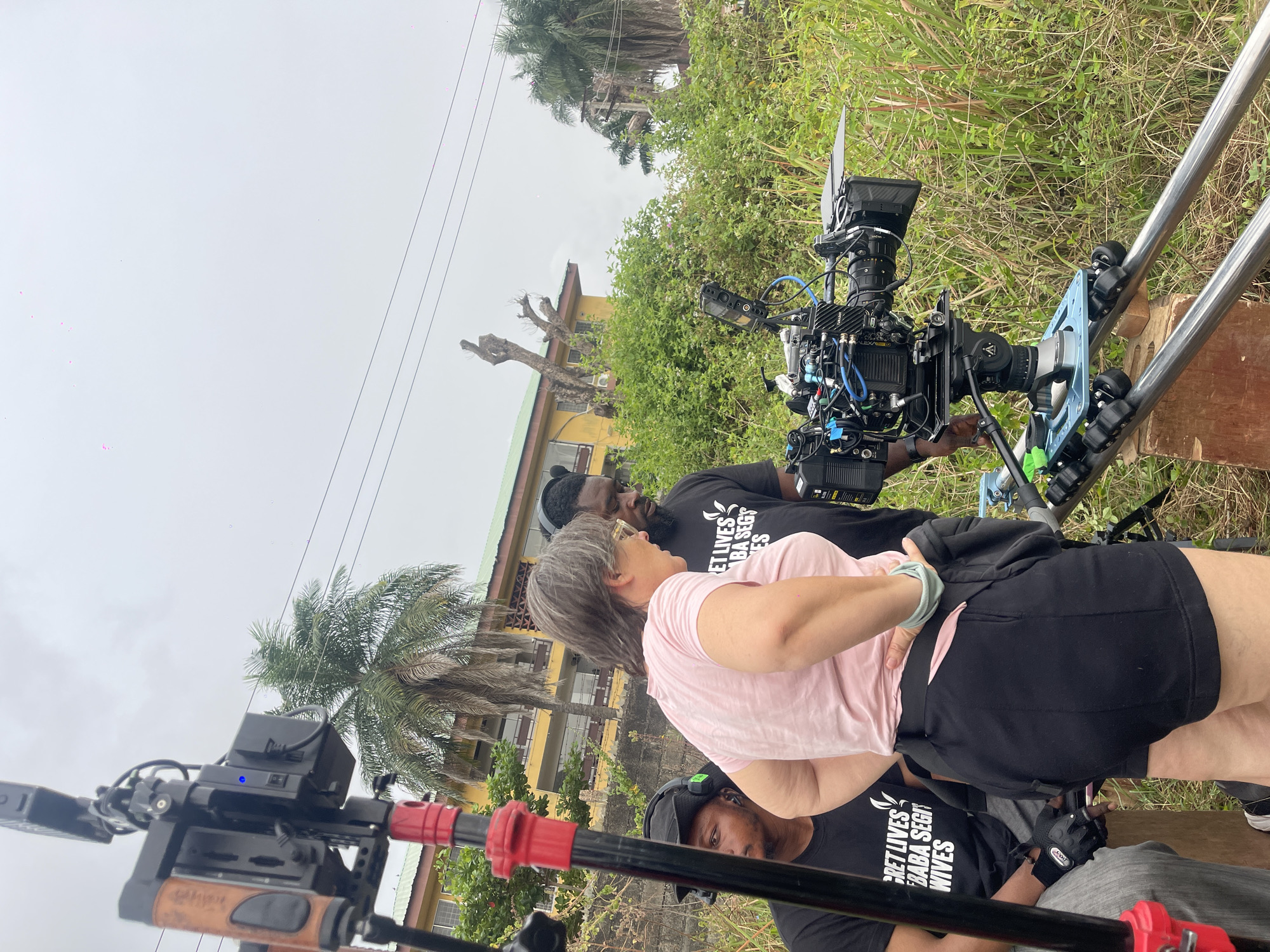 On location in Unknown, Unknown, a film crew operates a professional video camera mounted on rails. A woman with short grey hair and a pink shirt stands observing, flanked by two crew members wearing black shirts with white text. The background includes a yellow building, tall grass, and palm trees under an overcast sky. Overhead power lines are visible.