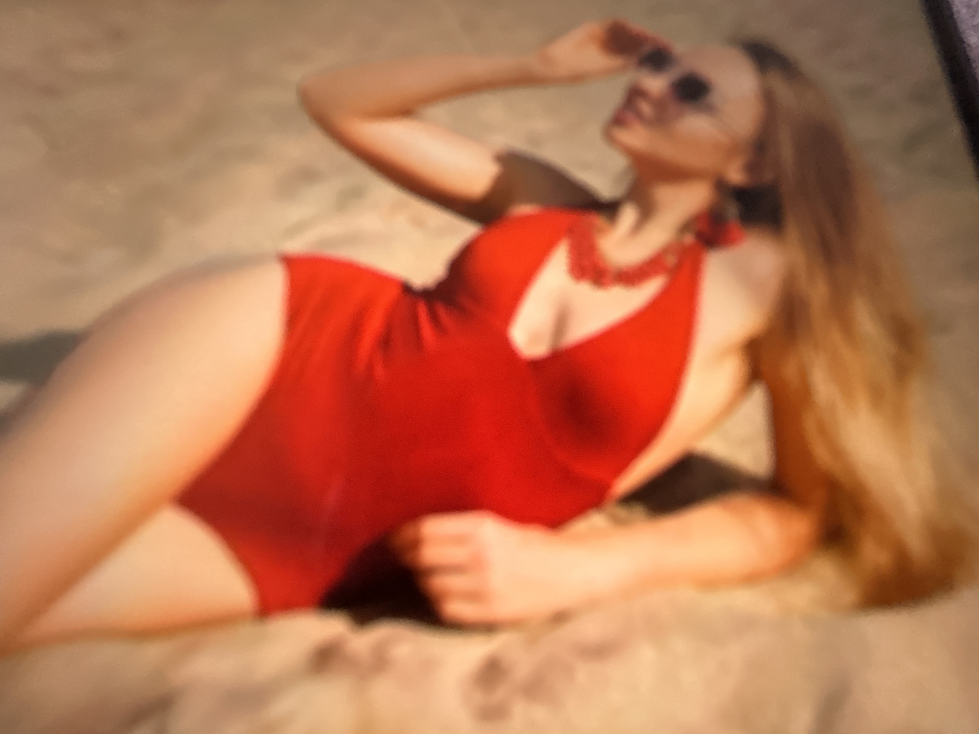 The image is a blurry, close-up shot of a person wearing a red swimsuit. The person is lying on their back with their legs in the air and their head tilted back. Their hair is long and blonde. They are wearing sunglasses and a beaded necklace. The background is a textured surface, possibly sand or fabric. The image appears to be taken outdoors, possibly on a beach or by a pool. There is no information in the image that would indicate a specific location in Gelendzhik, Russia.