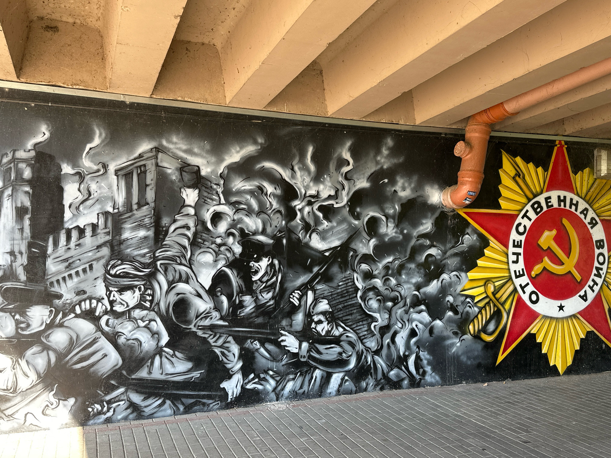 The image captures a mural on a concrete wall, likely located under a bridge or overpass. The mural is rendered in grayscale, depicting a scene of wartime struggle with stylized figures of soldiers and exploding buildings. Smoke billows across the composition, adding to the dramatic atmosphere. To the right of the mural, a large, red, five-pointed star with golden rays is prominently displayed. This star features a central circle with the hammer and sickle symbol, and text in Cyrillic script reads "ОТЕЧЕСТВЕННАЯ ВОЙНА" (Fatherland War). This imagery suggests the mural commemorates World War II, specifically the Eastern Front. The ground in front of the mural is paved with gray bricks. A light-colored pipe, possibly for drainage or utilities, runs along the ceiling above the mural. The overall scene is one of historical commemoration and artistic expression, with the red star and war imagery strongly indicating a connection to Soviet-era history and patriotic sentiment, common in public spaces in Belarus, including Brest.