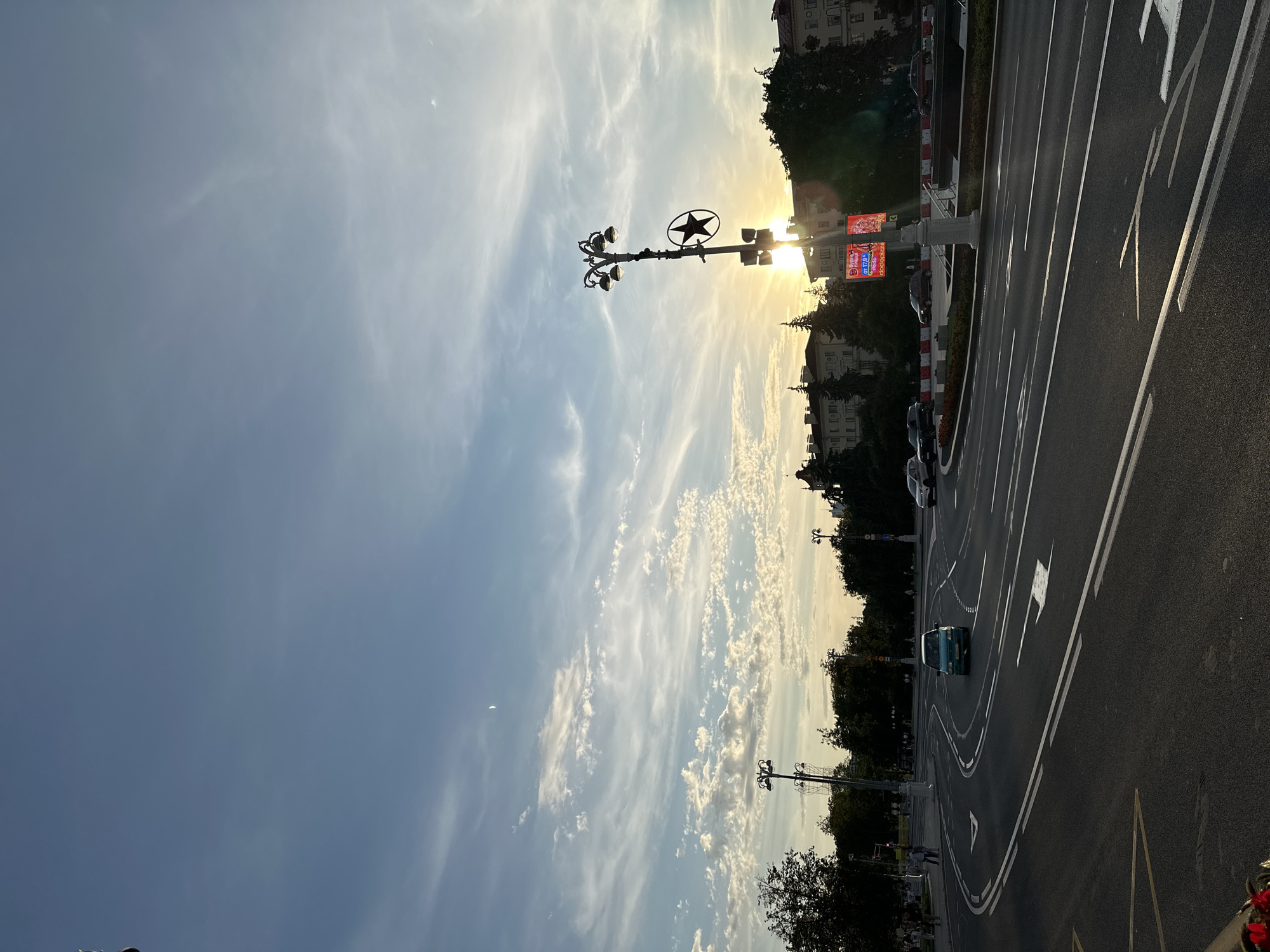 The image shows a street scene in Minsk, Belarus, during sunset. The sun is low in the sky, casting a warm glow and creating lens flare. A distinctive street lamp with a star emblem stands prominently in the foreground, silhouetted against the sun. The street itself is a multi-lane road with clear white lane markings. Several cars are visible on the road, including a turquoise minivan and a silver sedan. Pedestrians can be seen on a sidewalk in the background, and trees line the street. Buildings with neoclassical architectural styles are visible behind the trees. The sky is a gradient of blue and orange, with wispy clouds scattered across it. A crescent moon is also visible in the sky. The overall impression is of an urban environment at dusk, with a touch of historical charm.
