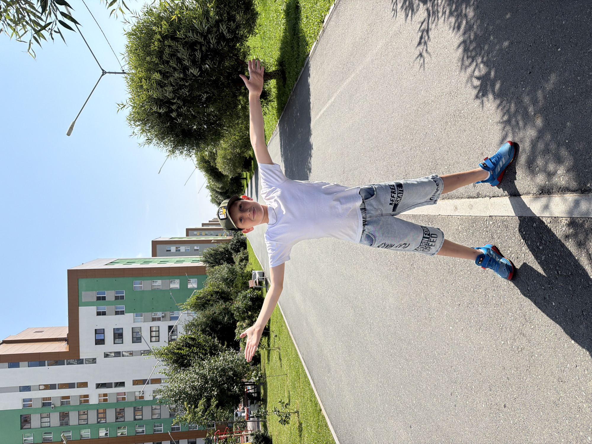 A young boy stands in the middle of an asphalt path, facing the camera with his arms outstretched to the sides. He is wearing a white t-shirt, light denim shorts with graphics, a dark baseball cap, and blue athletic shoes. Behind him, the path extends into the distance, flanked by green grass and trees on either side. In the background, multi-story residential buildings with a modern, colorful facade line the left side of the path, indicating an urban environment. Streetlights are visible along the path. The sky is clear and blue, suggesting it is a sunny day. This scene appears to be in a modern residential development, possibly in Yekaterinburg, Russia, given the architectural style and urban setting.