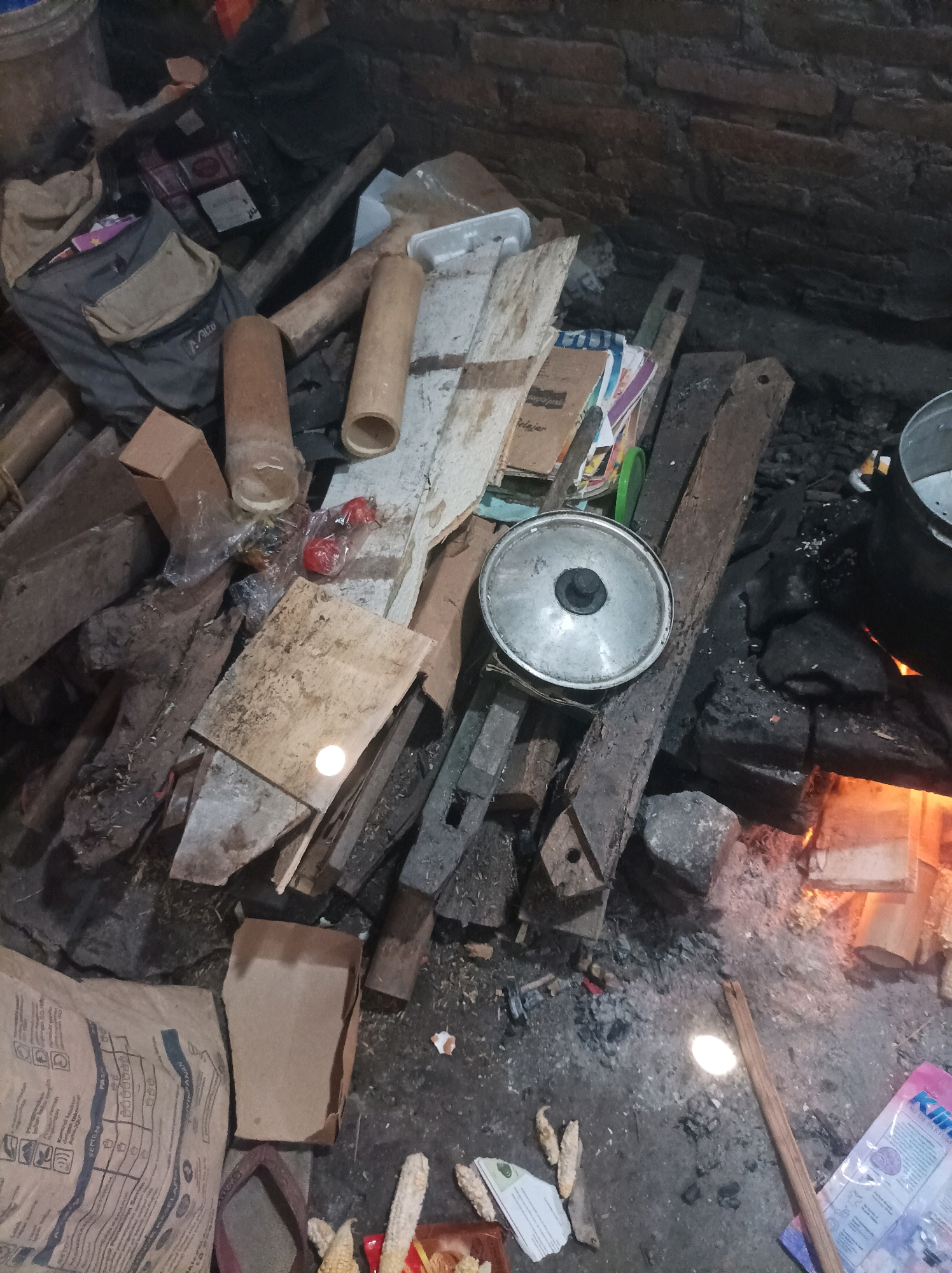 The image depicts an outdoor cooking area, likely a makeshift kitchen, with a fire pit actively burning in the lower right quadrant. The scene is cluttered with various objects, suggesting a humble or improvised living situation. In the foreground, a large, brown paper bag with printed text is visible, possibly containing ingredients or supplies. Adjacent to it, a small, overturned brown cardboard box and a red flip-flop are present. Several pieces of dried corn on the cob are scattered on the ground.
The central area of the image is dominated by a collection of wooden planks and debris, some appearing old and weathered. A silver pot lid with a black handle rests on top of some of these wooden pieces. To the left, there are cylindrical objects, one appearing to be made of bamboo and another possibly a cardboard tube. A gray backpack with tan accents is also visible in the upper left, alongside other assorted items and what appear to be plastic packaging.
In the background, a rough brick wall forms the backdrop. The fire pit is filled with glowing embers and burning wood, with a large black pot positioned over it, indicating that cooking is in progress. The ground is uneven and appears to be composed of dirt and ash. The overall impression is one of a functional, albeit unkempt, outdoor cooking space within the village of Canga'an, Indonesia. No individuals are visible in the image.