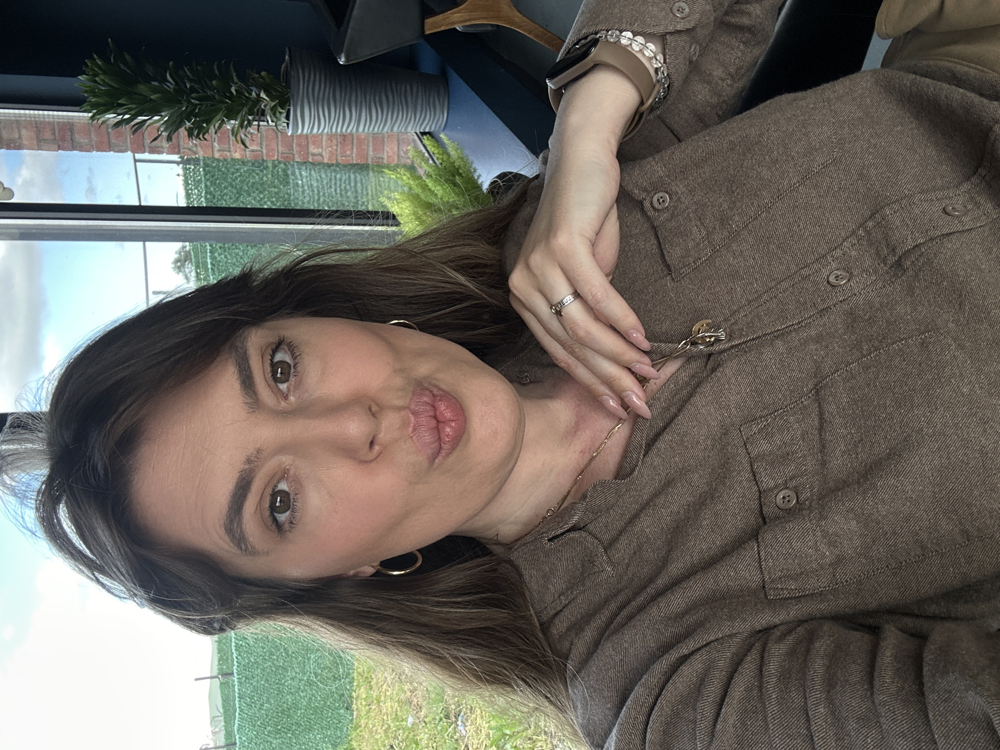 A woman is posing indoors for a selfie. She is looking at the camera with puckered lips. She has long brown hair, gold hoop earrings, a gold chain necklace, and is wearing a brown button-down shirt. She also wears a tan watch and a ring on her left hand. Outside the window behind her, there is grass, fencing, and trees. Brick buildings are visible in the background. The location in Marneuli, Georgia cannot be confirmed from the image.