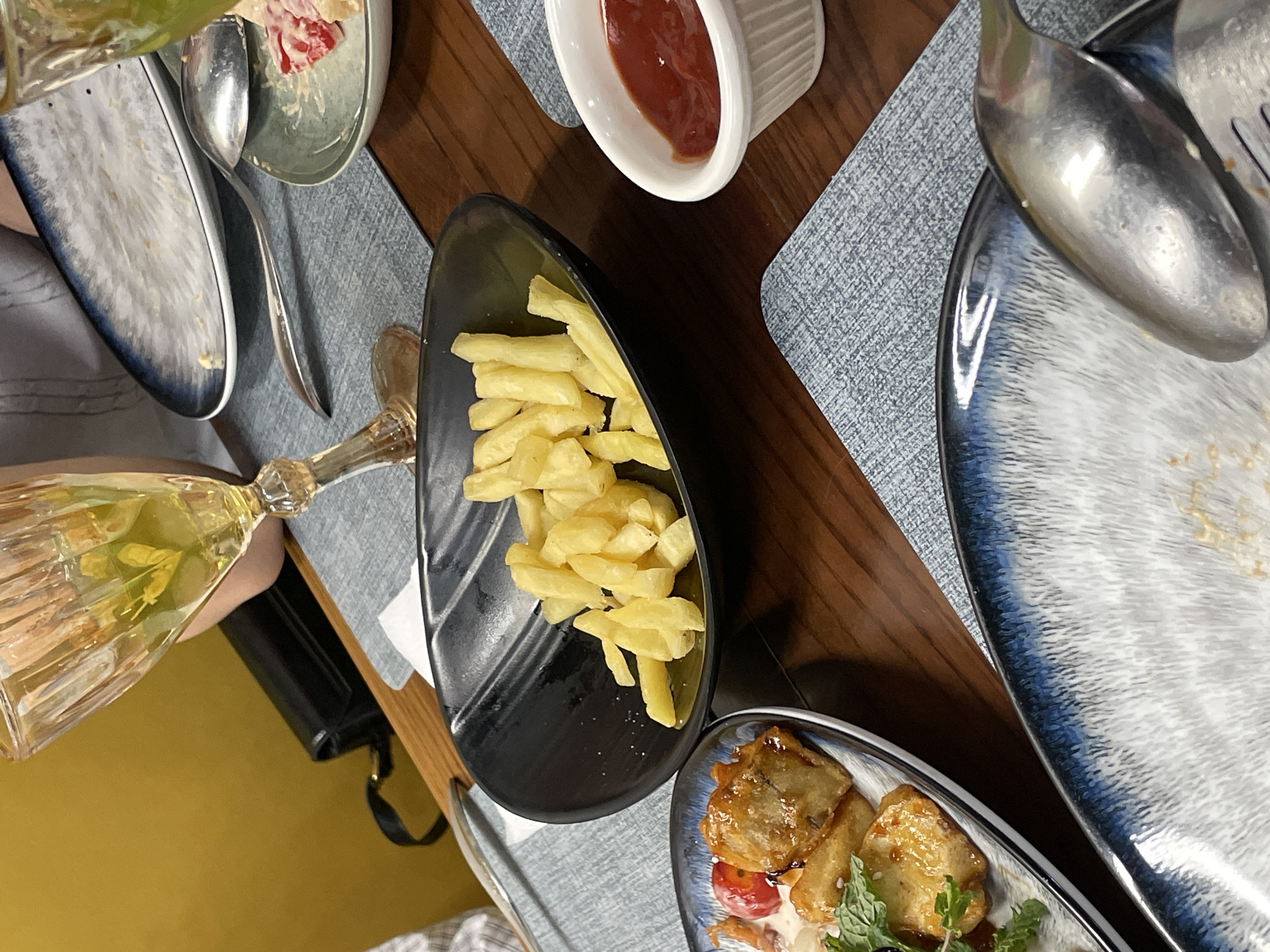 The image shows a close-up view of a wooden table at a restaurant, likely in Tashkent, Uzbekistan, laid with blue and white patterned placemats and various dishes. In the foreground, a black oval bowl filled with french fries sits prominently. To the right of the fries, a small white ramekin holds a dollop of red sauce, presumably ketchup. Further to the right, out of focus, is a white bowl containing what appears to be a salad with red pieces and creamy dressing, partially eaten. Behind the fries, a tall, stemmed glass with a yellow beverage and a sprig of greenery is visible, reflecting light. Another plate, partially visible in the upper left, displays food and a black handbag rests behind it. In the lower right corner, a spoon and fork are resting on a plate with a similar blue and white pattern. The overall scene suggests a dining experience in a casual to mid-range establishment.