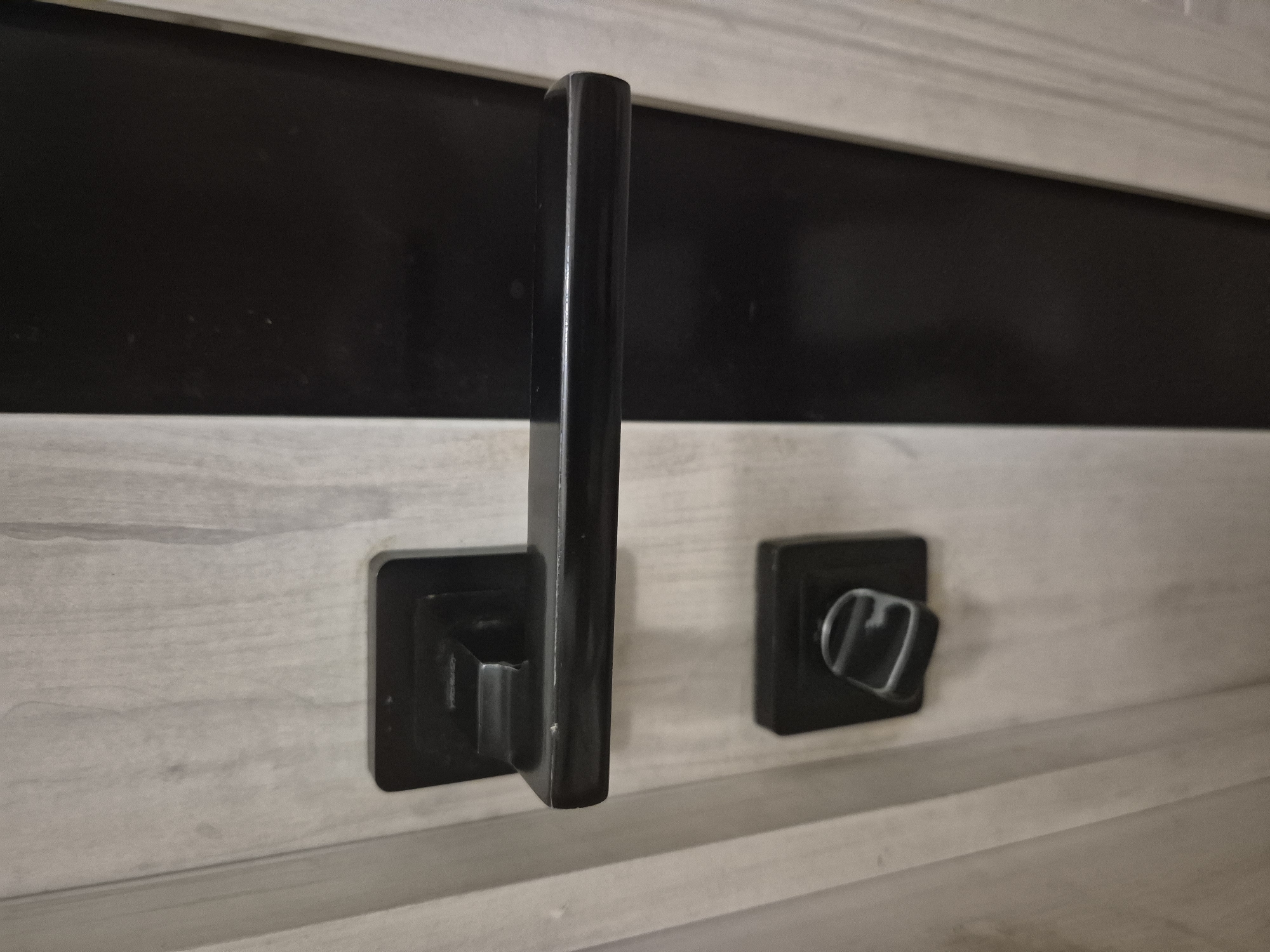The image shows a close-up of a black door handle and lock mechanism on a light-colored wooden door. The door has a vertical dark panel on the right side. The handle is rectangular with a horizontal bar, and the lock is a smaller square mechanism with a turnable knob. The scene appears to be indoors, likely a room within a building. There are no people or animals visible in the image. The lighting suggests an indoor setting, possibly with ambient or artificial light. The overall impression is of a functional, modern door hardware. There is no information in the image to suggest a specific location in Astana, Kazakhstan.