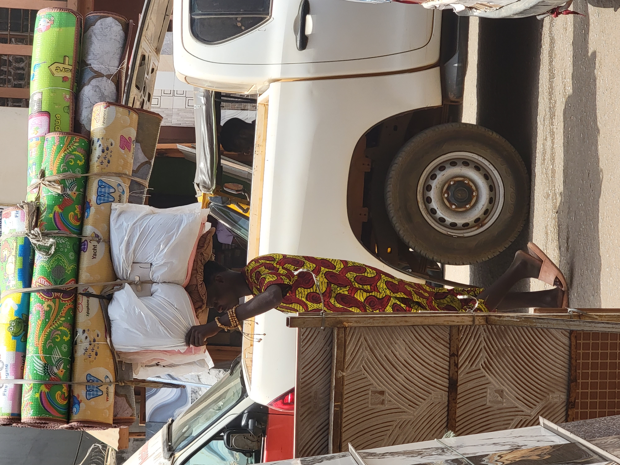 In Maiburiji, Nigeria, a white vehicle is parked on a paved street. A person wearing a colorful patterned dress and sandals is leaning over a rusty, patterned metal barrier next to the vehicle's tire. The vehicle appears to be loaded with a stack of items including rolled mats, white bags, and other miscellaneous goods. Another vehicle is partially visible in the foreground. The scene is bathed in sunlight.