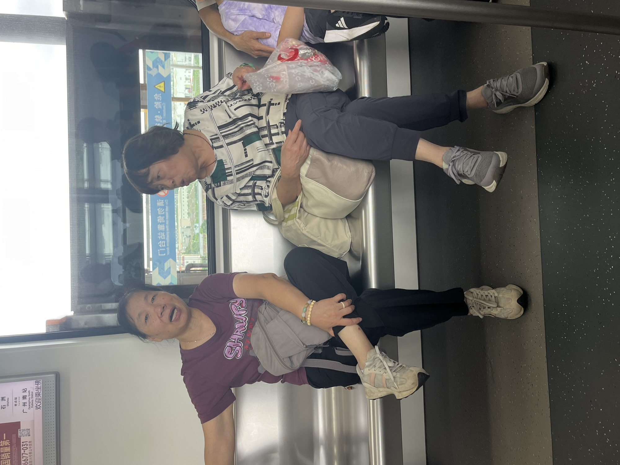 Two women are sitting on a metal bench inside a subway car. The woman on the left is wearing a dark purple t-shirt with white text that says "SHNVF", black pants, and light-colored athletic shoes. She has a gray backpack on her lap and is wearing bracelets on her left wrist. She is looking towards the camera with her mouth open. The woman on the right is wearing a black and white patterned shirt, dark pants, and gray athletic shoes. She is holding a clear plastic bag with a red object inside. She is looking to her left with her mouth partially open. The subway car has large windows that show a blurred view of buildings and sky. A sign above the window on the left displays Chinese characters and the text "Guangzhou South Railway Station" and "欢迎乘伴" (Welcome aboard). This indicates the location is likely within the Guangzhou Metro system, possibly serving the Shunde District in Foshan city, Guangdong Province, China, given the context of Guangzhou South Railway Station's proximity to Shunde.