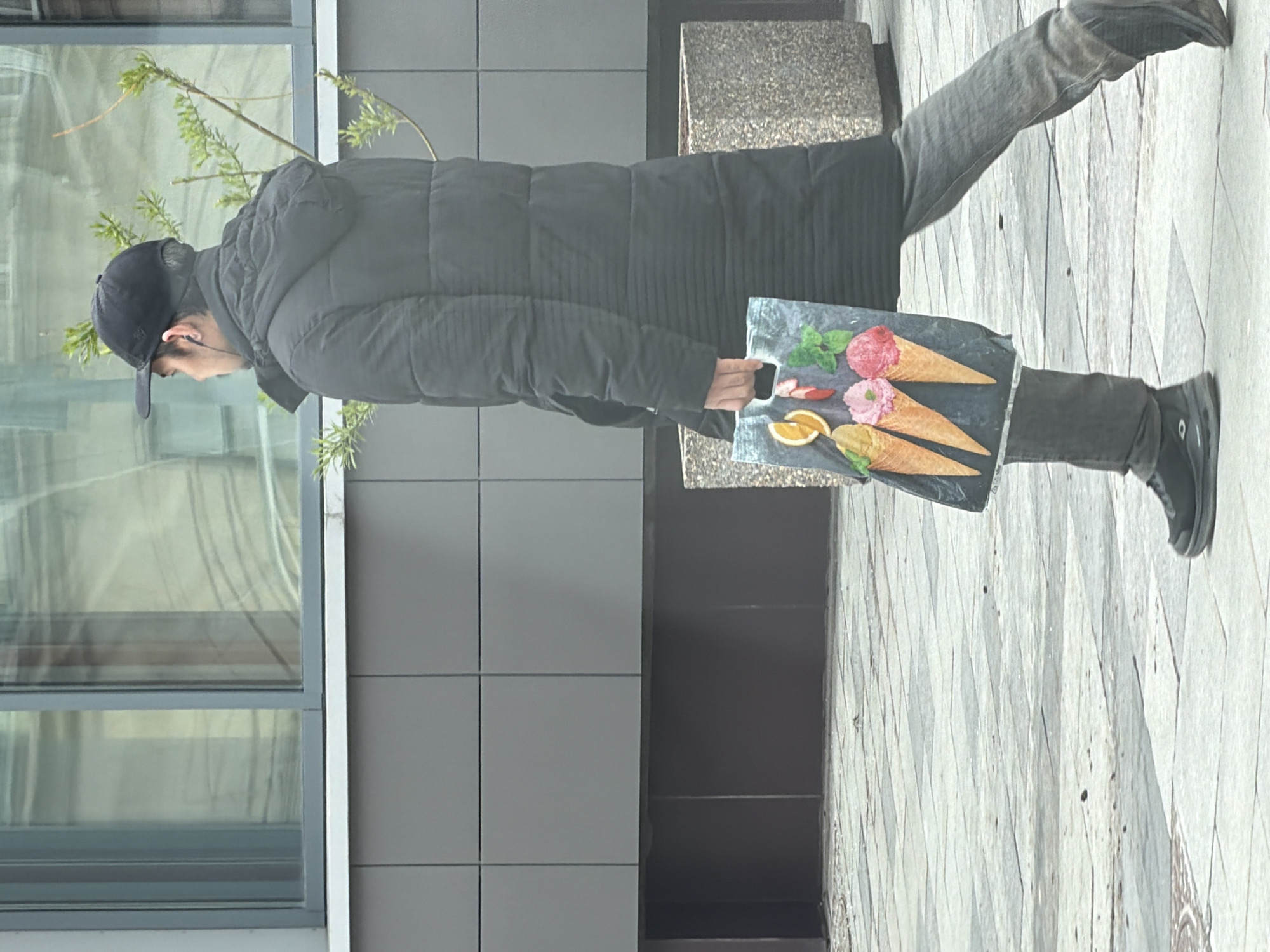 A person is depicted walking outdoors in what appears to be an urban setting, likely a street or sidewalk in Moscow, Russia. The individual, seen from a side profile, is wearing a black baseball cap, a dark, puffy winter coat, dark jeans, and black shoes. They are holding a flat, dark rectangular object, possibly a tote bag or a display board, which features vibrant, realistic illustrations of three ice cream cones. Two of the ice cream scoops are pink (strawberry or raspberry flavored), and one is a pale yellow/light brown (possibly vanilla or mango), all nestled in waffle cones. The illustrations also include garnishes such as sliced strawberries, orange wedges, and green mint leaves.
The background shows a modern building with large glass windows reflecting the surroundings, and dark gray rectangular panels on its facade. To the right of the person, there is a gray concrete or stone planter. Small green branches or foliage are visible behind the person's head, suggesting budding plants or bushes. The ground is a light-colored pavement with a distinctive geometric, diamond-shaped pattern. The scene is illuminated by daylight, and there are no strong shadows, indicating an overcast day or indirect light. No text is visible in the image.