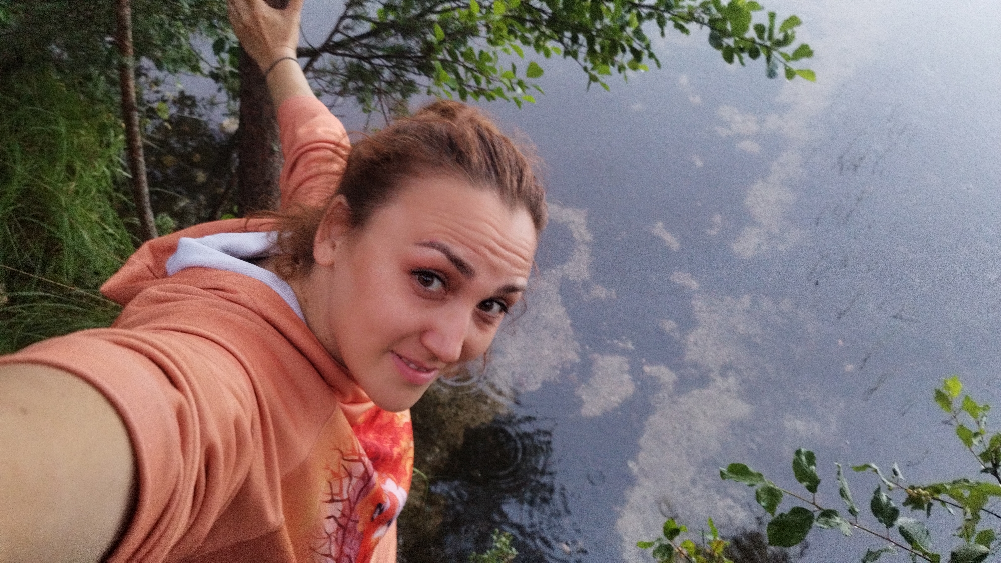 A high-angle medium shot shows a young woman with reddish-brown hair in a loose bun, facing the camera and smiling. She is wearing a coral-colored hooded sweatshirt with a white lining at the neck. The woman is outdoors, with her left arm extended as if taking a selfie. To her right, a body of water, likely a lake or river, fills the majority of the frame, with reflections of the sky and surrounding vegetation visible on its surface. To the left of the woman, tall green grass and a thin tree trunk are visible. Green leaves from a tree branch hang into the upper right corner of the frame, partially obscuring the water. The overall scene suggests a natural, outdoor setting.