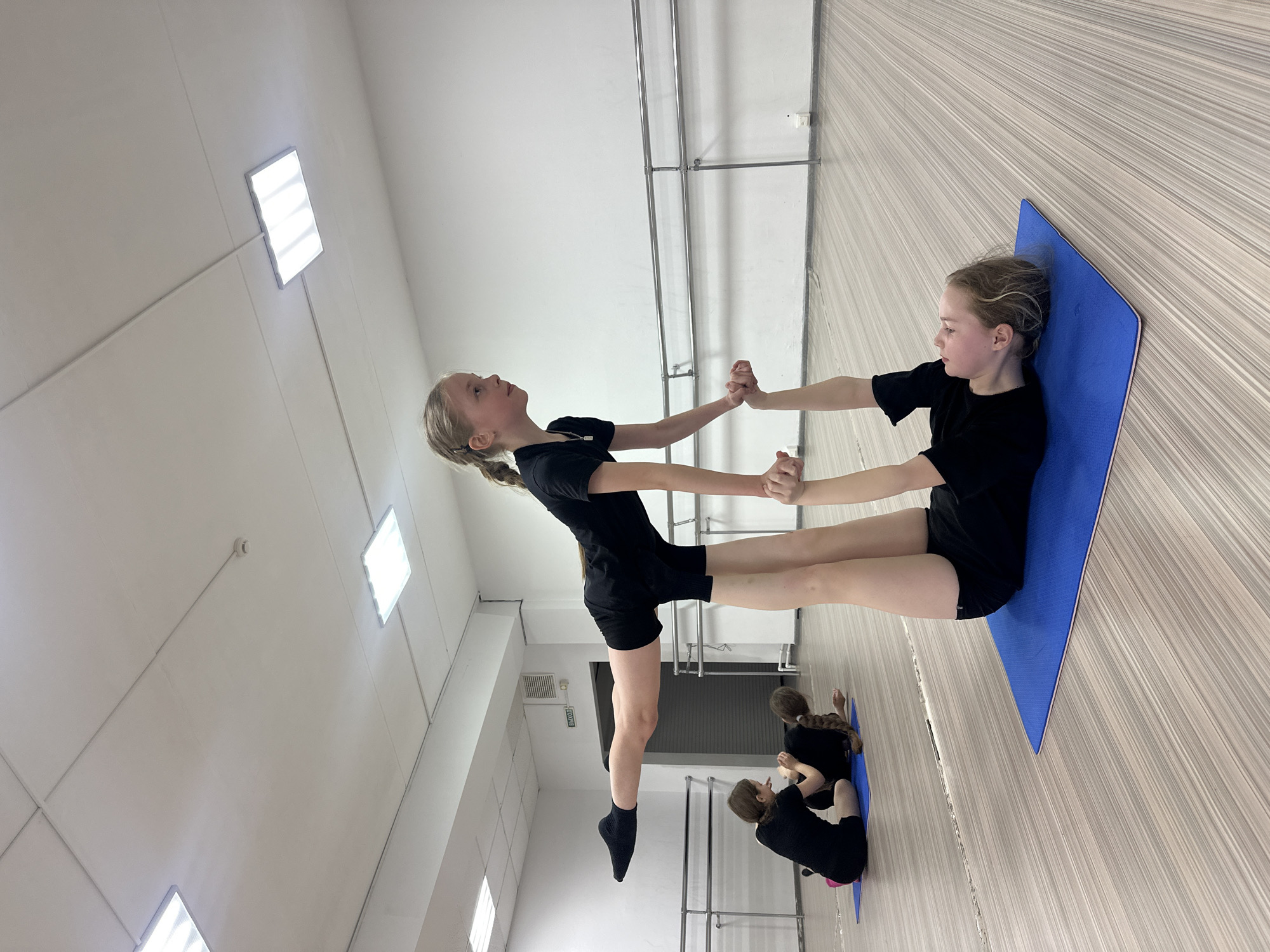The image displays an indoor scene within what appears to be a gymnastics or dance studio, likely located in Votkinsk, Russia. Four young girls, all dressed in black athletic attire (t-shirts/leotards and shorts), are present.
In the foreground, two girls are performing a partner acrobatic or balance pose. One girl is lying on her back on a blue exercise mat, holding the hands of the second girl. The second girl is standing and balancing, with her legs extended upwards, maintaining a poised, elevated posture. Her left foot is bare and wearing a black sock, while the right foot is not visible. Both girls exhibit focused expressions.
In the background, two additional girls are seated on separate blue mats, seemingly resting or observing the main activity. The studio features light-colored walls and a light-colored floor with a distinct wood-grain pattern. Metal bars, resembling ballet barres or gym equipment, are mounted along one wall. Overhead, several rectangular fluorescent light fixtures illuminate the space, and a white fire alarm or smoke detector is visible on the ceiling. A white sign with the Russian word "ВЫХОД" (EXIT) is visible on a door or panel in the background. The overall impression is of a dedicated training session for gymnastics or dance.