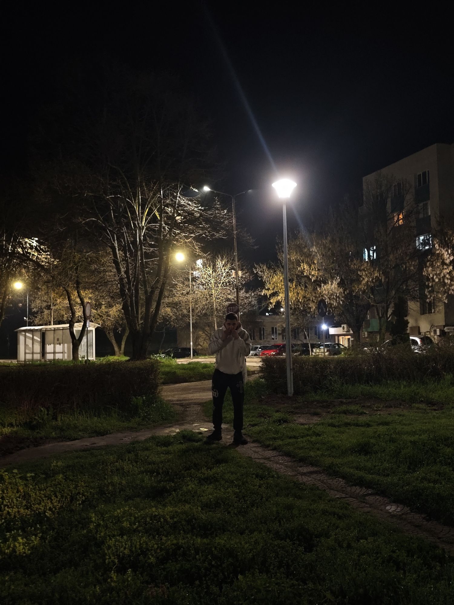 A medium shot captures a man standing on a grassy patch in Kozloduy, Bulgaria. He is dressed in casual attire, including a light-colored hoodie, dark pants, and black shoes. He appears to be holding something up to his face. The scene is lit by several street lights, creating a contrast between illuminated areas and shadows. There is a bus stop and a building in the background. The scene takes place at night.