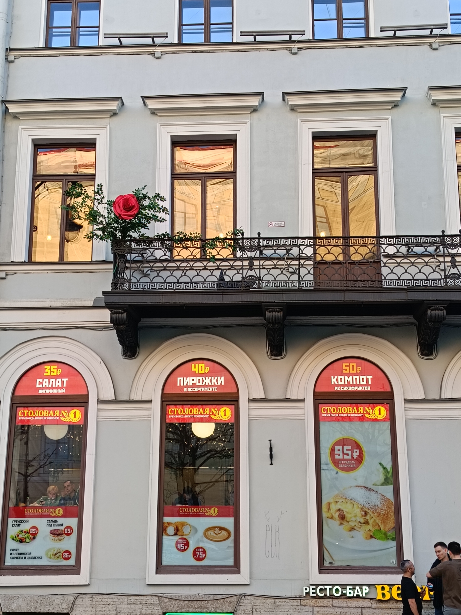 The image shows the facade of a building in Saint Petersburg, Russia. The building has two stories and is painted light blue with white trim. The upper story has windows with dark wood frames and a balcony with an ornate black metal railing. A large red rose bush is growing on the balcony. The lower story has arched windows with red signs advertising food and drinks. The signs are in Russian and offer items such as salad (35₽), pirozhki (40₽), and kompot (50₽). There are also posters visible within the windows advertising specific dishes. A sign at the bottom reads "РЕСТО-БАР BE". A group of four men stand in front of the building on the right side.