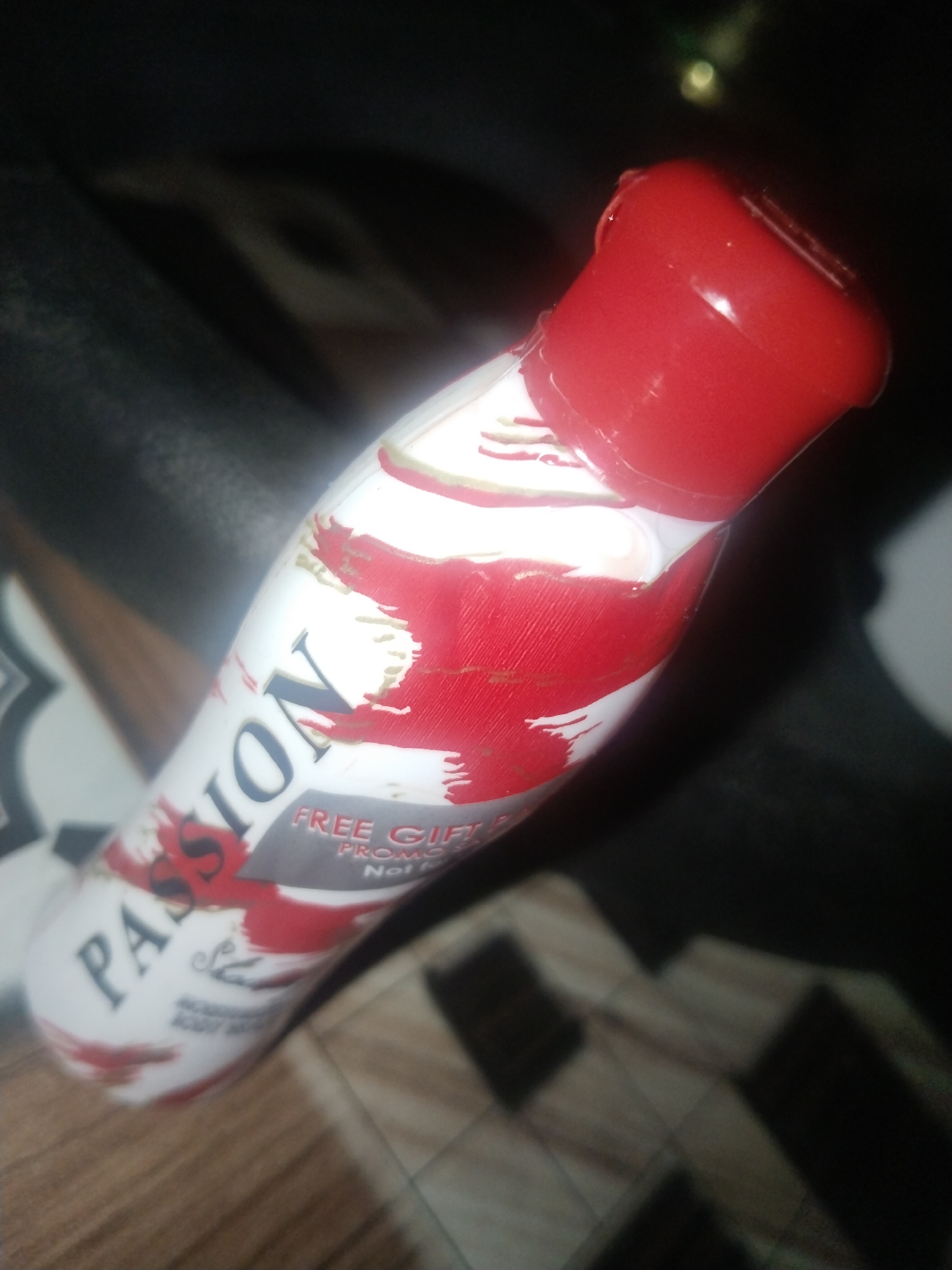 Here's a description of the image:
The image shows a bottle of "Passion" body wash with a red cap. The bottle is white with red abstract designs. Text on the bottle indicates a "Free Gift" promotion. The bottle rests on a surface with a checkered pattern. The location is unknown.