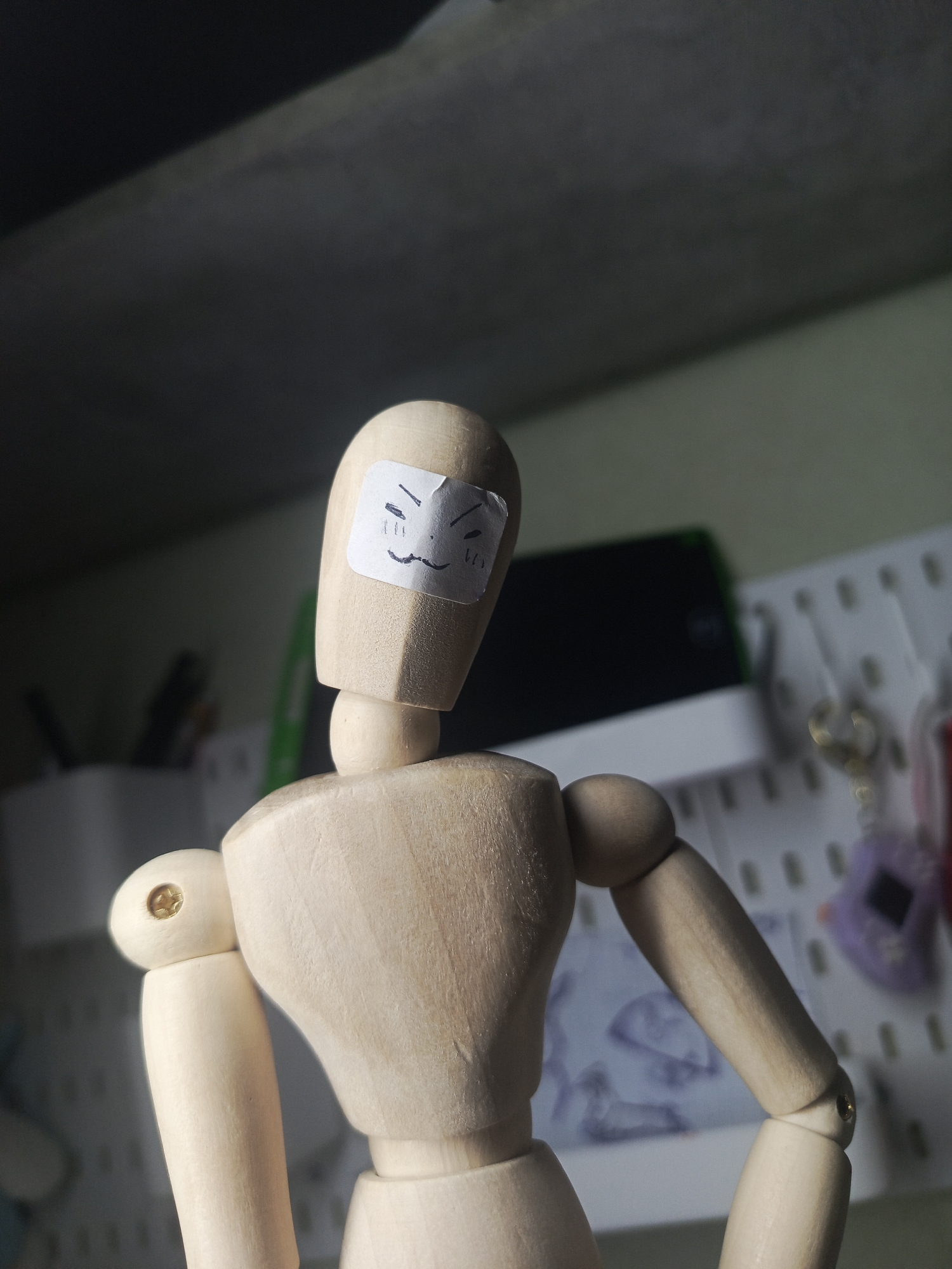 A close-up shot of a wooden mannequin with an angry cartoon face taped to its head. The mannequin is in a room with a shelf in the background. On the shelf is a black object, a white container with pens, and a pegboard with various small objects hanging on it, including a purple keychain. The mannequin's right arm is bent and resting on its hip.