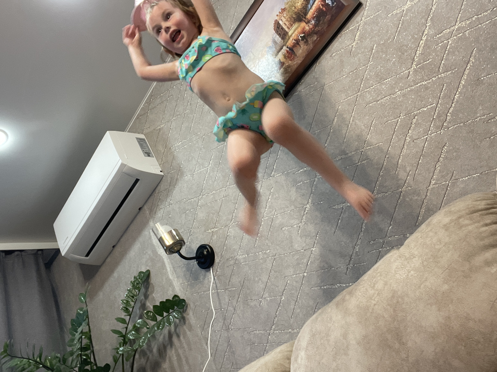 The media file depicts a young girl in a bikini mid-jump in an indoor setting. She is wearing a light blue bikini with a fruit pattern and a pink headband. Her arms are outstretched, and her mouth is open in a joyful expression. The background features a grey, textured wallpaper with a geometric pattern, an air conditioning unit mounted on the wall, a small wall lamp, and a framed picture hanging above. To the right, a portion of a beige sofa is visible. The lighting suggests it could be daytime, with light coming from an unseen source. The overall scene conveys a sense of playful energy and childhood fun. There is no visible text in the image.