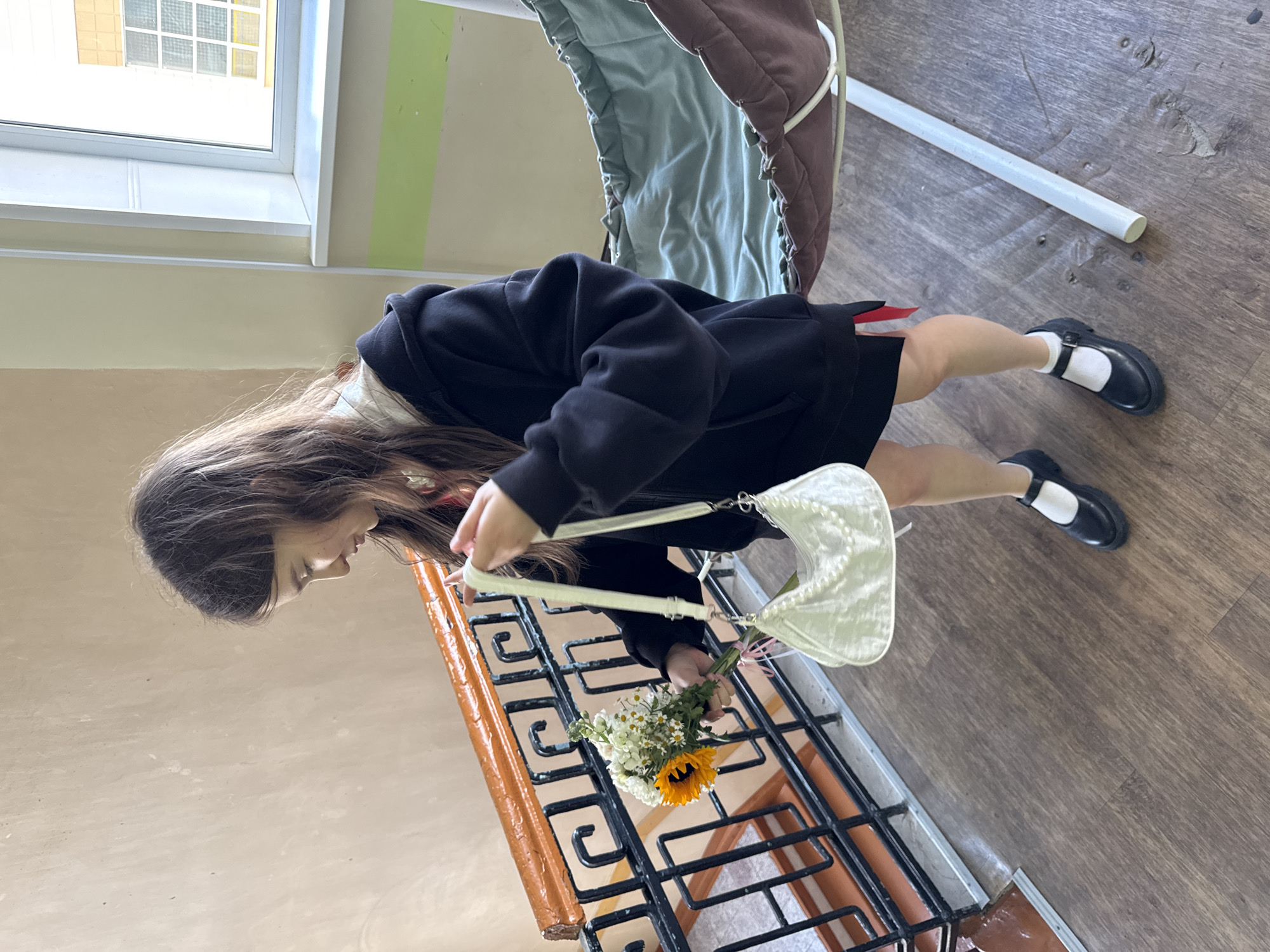 A young woman stands on a staircase in a building. She wears a black hoodie, a short black skirt, white socks, and black Mary Jane shoes. She is carrying a white purse and a bouquet of flowers containing a sunflower. The staircase has an orange and black railing. The environment suggests an institutional building with painted walls and a window with multiple panes. The scene is in Ivanovo, Russia.