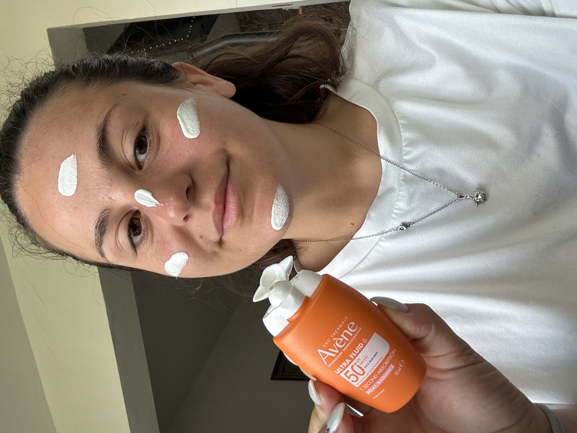 A young woman is applying sunscreen to her face. She is holding an orange bottle of Avène Ultra Fluid SPF 50 sunscreen in her left hand. Several white dots of sunscreen are visible on her forehead, cheeks, and chin. The woman is wearing a white t-shirt and a silver necklace with a star pendant. The background is blurred, but appears to be an indoor setting with a pale green wall and a dark gray cabinet. There is no visible indication of the location being Plovdiv, Bulgaria.