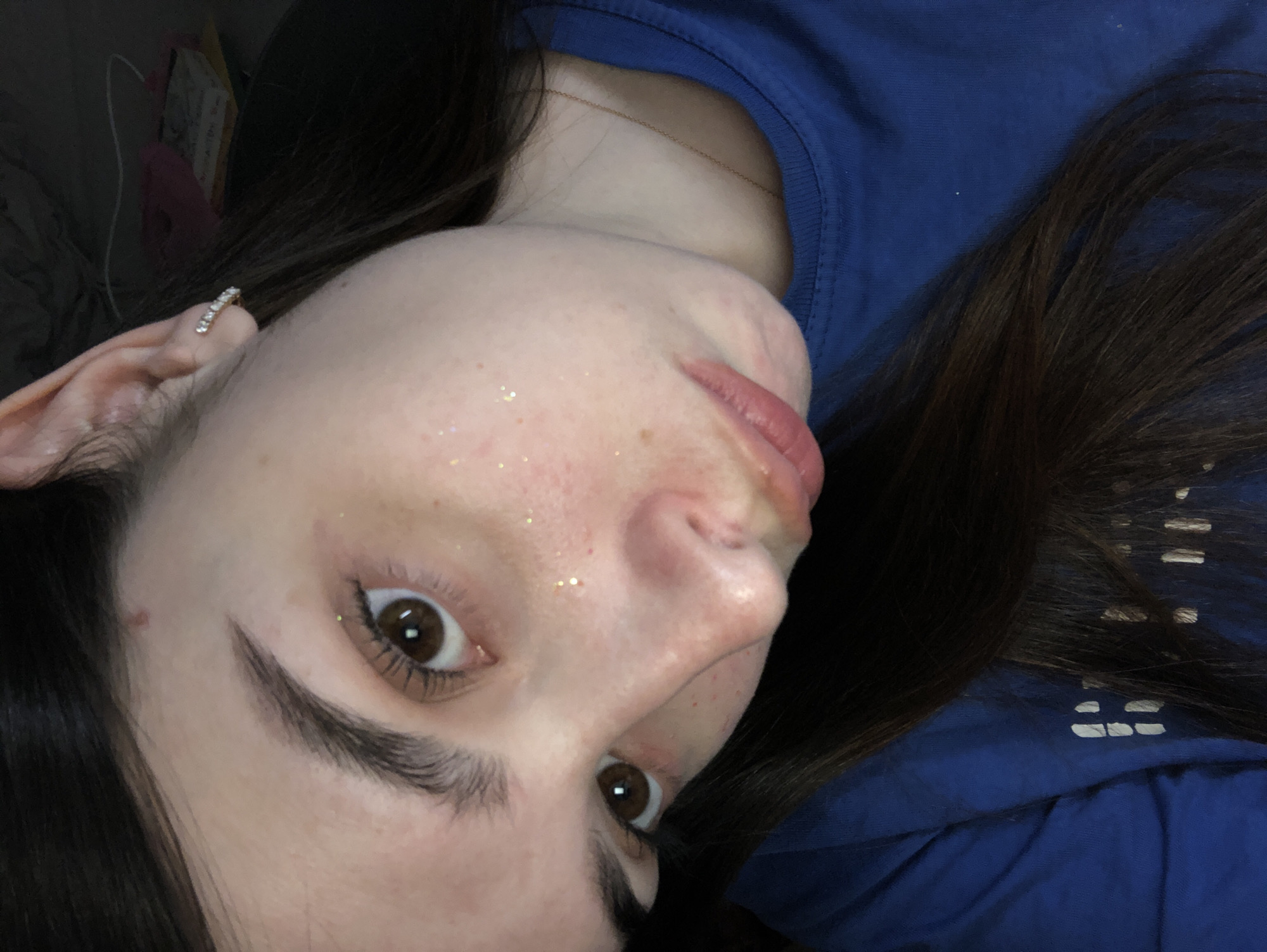 A close-up, angled selfie shows a young woman with brown hair and brown eyes. She is wearing a blue shirt and a small, jeweled earring. Her skin has a light dusting of glitter. A thin gold necklace is visible. The background is dark and indistinct, with a glimpse of a partially visible pink and white object. The image offers no information to suggest the location is Noviy, Russia.