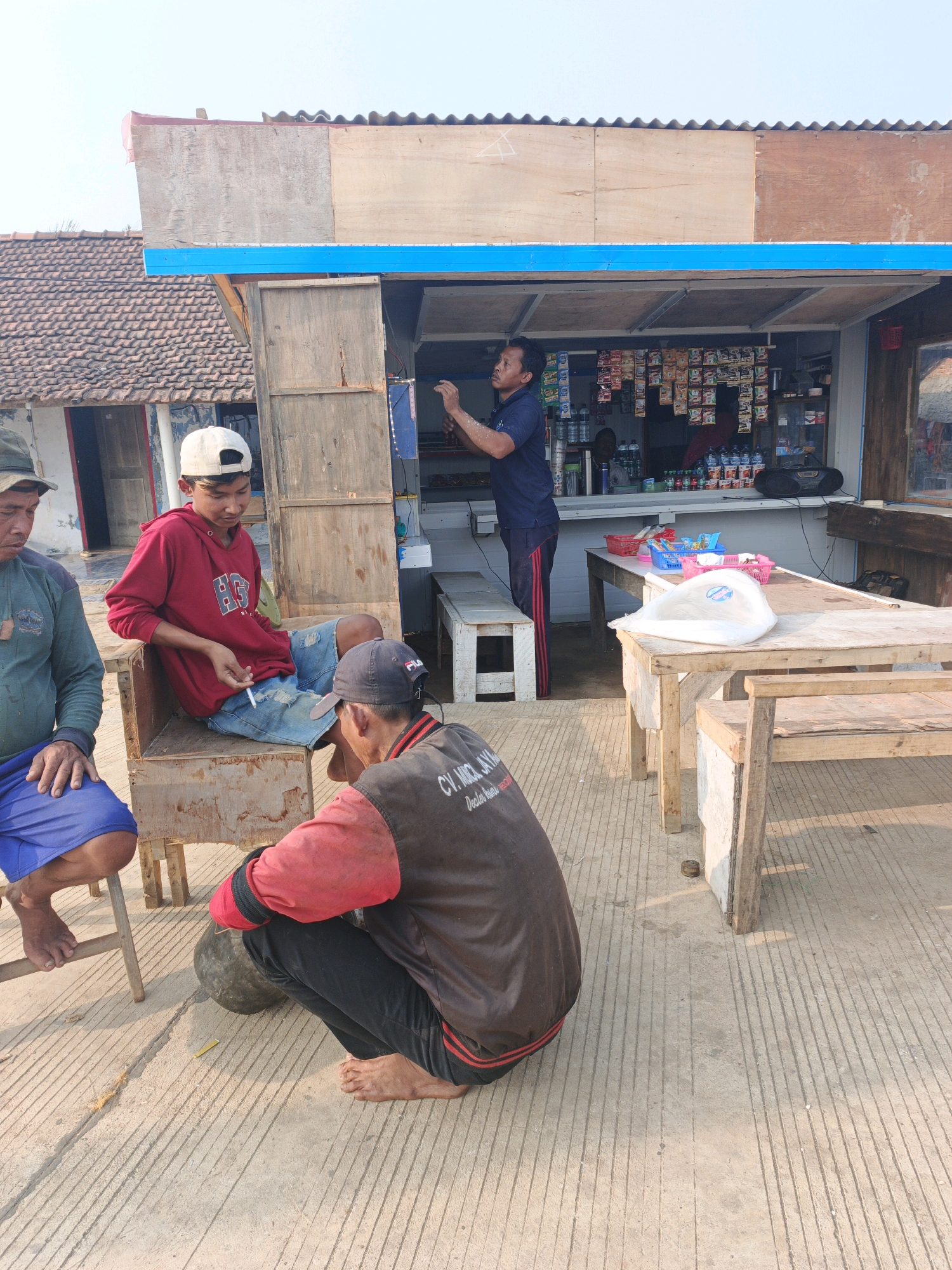 The image depicts an outdoor scene in Unknown, Indonesia, featuring a small roadside stall or kiosk. In the foreground, three men are present. One man, wearing a red hooded sweatshirt and denim shorts, is seated on a wooden chair and smoking a cigarette. Another man, in a dark jacket with red sleeves and a black baseball cap, is crouched down, facing away from the viewer. A third man, partially visible on the left, is wearing a green long-sleeved shirt and blue shorts.
The stall behind them is constructed with a wooden frame and has shelves stocked with various packaged goods, likely snacks and drinks. A blue awning extends over the front of the stall. In front of the stall, there are two wooden tables with benches. One table has several plastic baskets and a white plastic bag on it. The ground is paved with concrete that has visible grooved lines. The background shows a residential structure with a tiled roof and a clear, light-colored sky. The overall impression is of a casual, everyday scene at a local vendor.