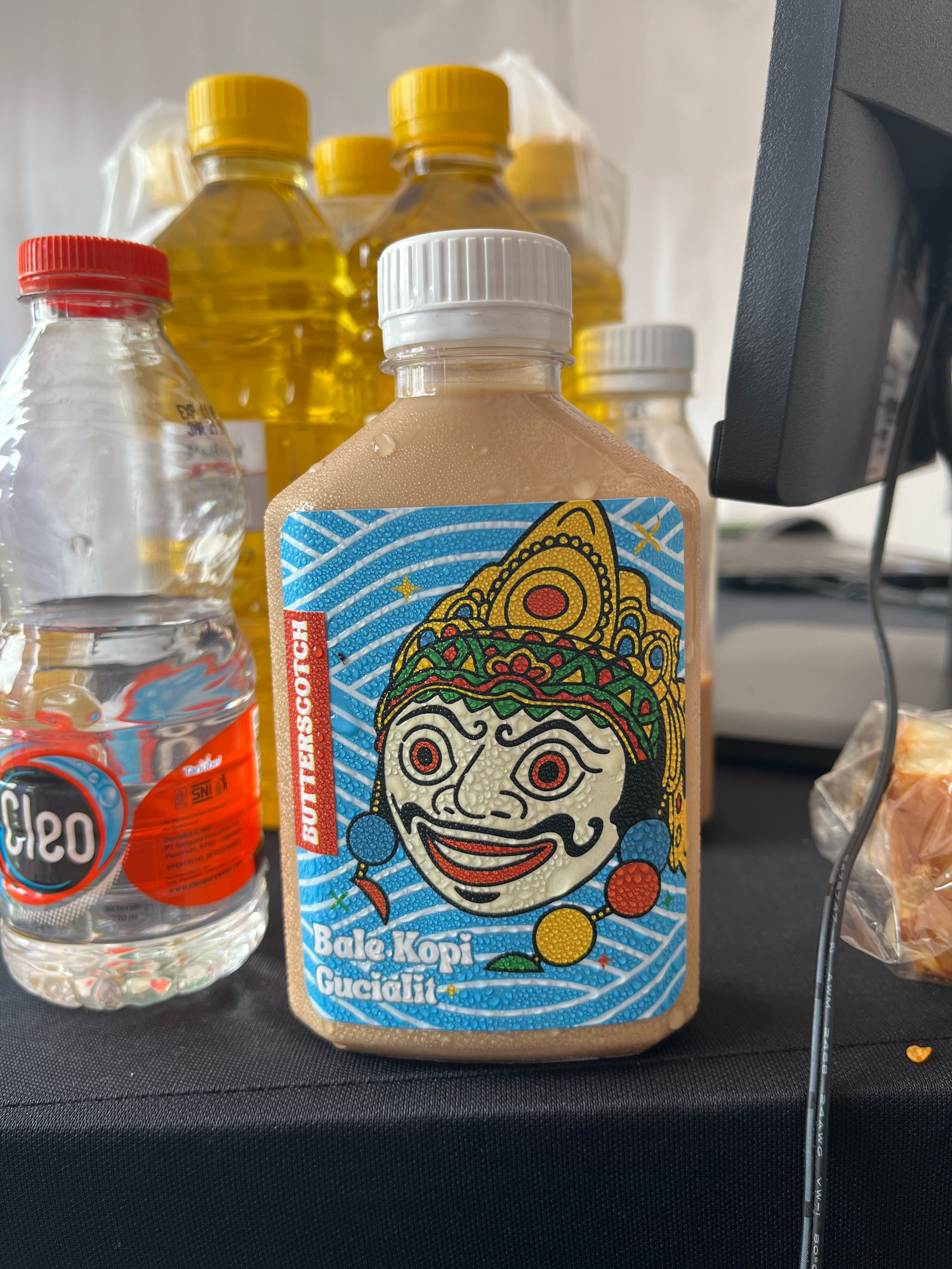 A close-up, eye-level shot captures several bottles on a black surface. In the foreground, a clear plastic bottle of Cleo brand drinking water is visible, its red cap slightly out of focus. To its right, a bottle with a white cap contains a light brown liquid, likely a beverage, with condensation on its surface. This bottle features a vibrant label with a stylized depiction of a traditional Indonesian puppet character. Text on the label reads "BUTTERSCOTCH" vertically on the left and "Bale Kopi Gucialit" horizontally on the bottom. Behind these two bottles, several larger plastic bottles with yellow caps are present, filled with a golden liquid, possibly cooking oil or a similar beverage. The background is a plain, light-colored wall. A black computer monitor or similar electronic device is partially visible on the right side of the frame. A plastic-wrapped food item, possibly bread, is also present on the far right. The lighting suggests an indoor setting. The presence of "Gucialit" on the beverage label suggests the location may be near or related to Gucialit, a subdistrict in the Lumajang Regency of East Java, Indonesia.