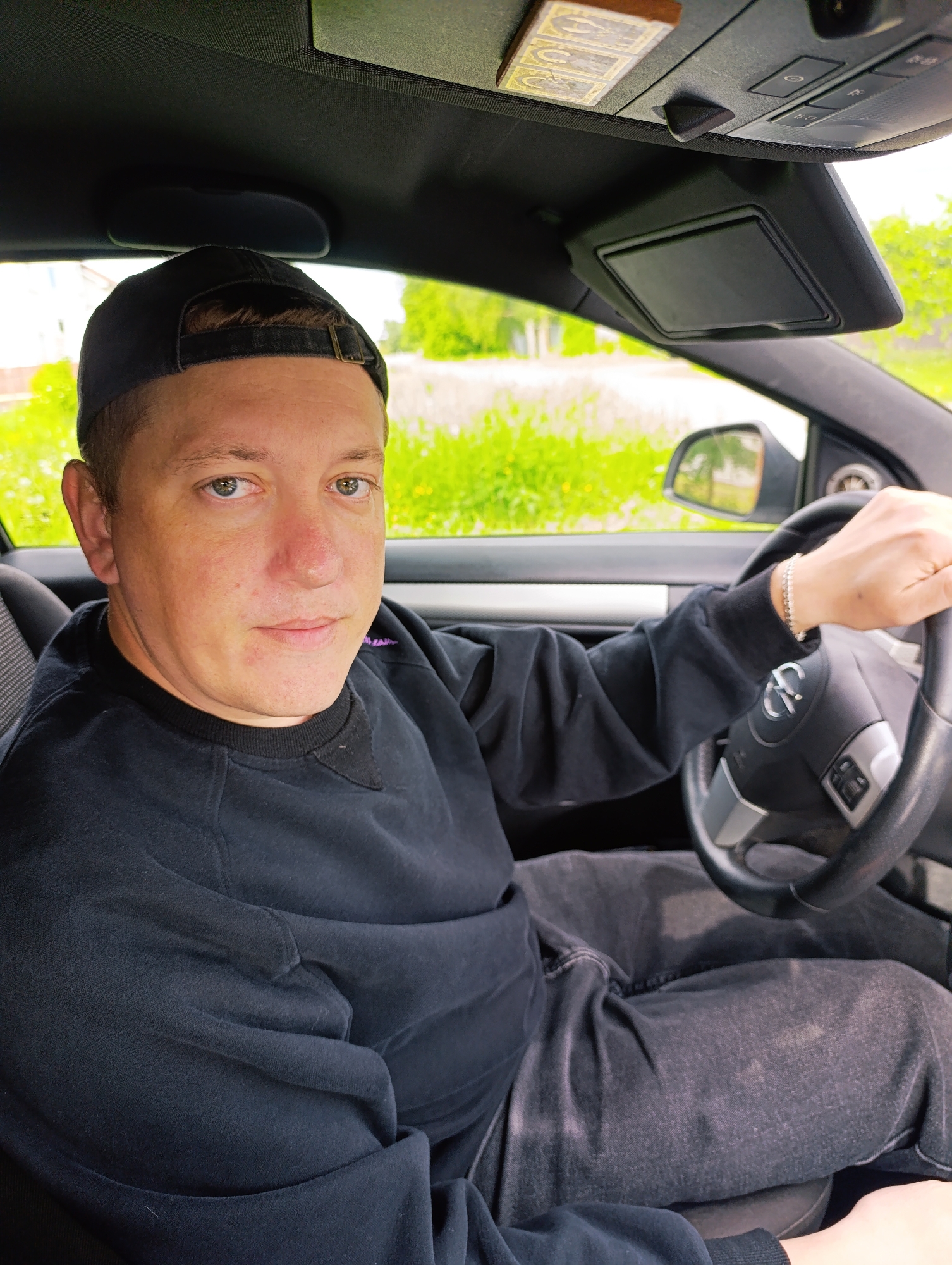A Caucasian male wearing a black baseball cap, a black long-sleeve shirt, and black jeans is sitting in the driver's seat of a car. He is looking directly at the camera with a neutral expression. His right hand is on the steering wheel, and he is wearing a bracelet on his right wrist. The car's interior is visible, including the dashboard, steering wheel, and sun visor. Through the windshield, greenery is visible, suggesting an outdoor setting. The location appears to be in the city of Unknown, Unknown.