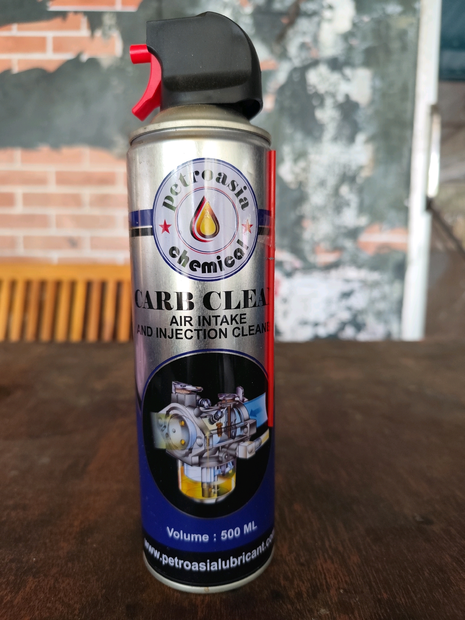 A silver aerosol can of "PETROASIA CHEMICAL CARB CLEANER AIR INTAKE AND INJECTION CLEANER" stands upright on a wooden surface. The can has a black spray nozzle with a red trigger. The label features a circular logo with the words "petroasia chemical" and a graphic of oil dripping into a droplet. Below the logo, "CARB CLEANER" is prominently displayed in large, black letters, followed by "AIR INTAKE AND INJECTION CLEANER" in smaller text. A diagram of a carburetor is visible on the lower part of the can. Text at the bottom reads "Volume: 500 ML" and "www.petroasialubricant.com". In the background, a blurred brick wall and wooden slatted furniture are visible. The scene is likely indoors or in a sheltered outdoor area in Palangka Raya, Indonesia, given the typical environmental conditions.