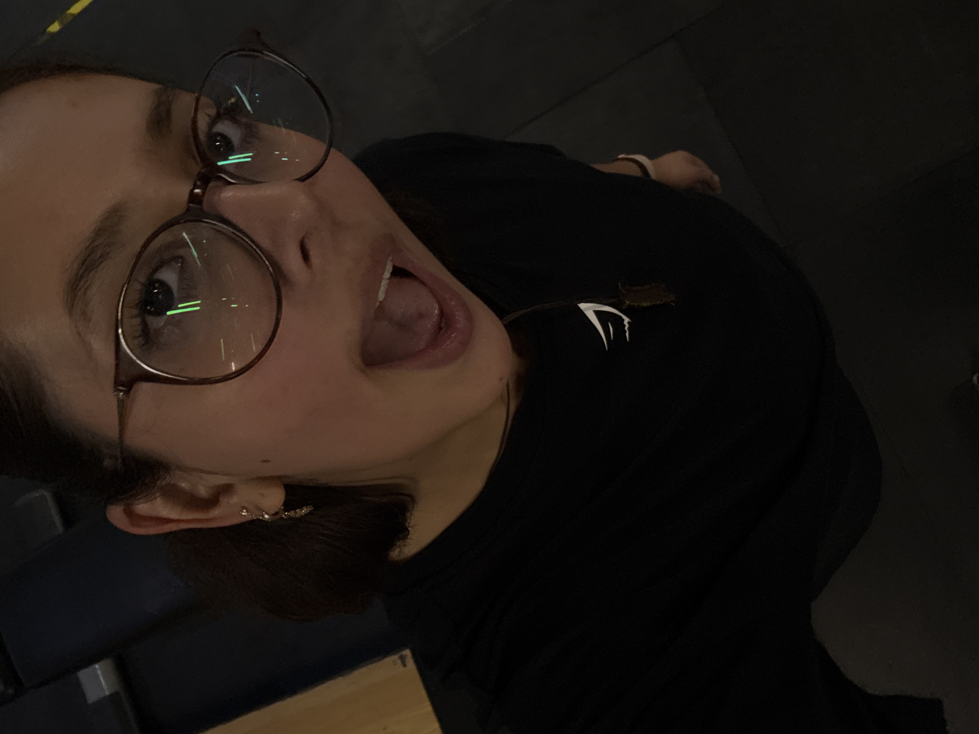 A woman with brown hair pulled back is wearing round, brown-framed glasses and is looking up at the camera with her mouth open. She has an earring in her left ear. She is wearing a black t-shirt with a white logo on the chest. The background is dark and blurry, with a hint of furniture visible on the left side of the frame. The location is San Andrés Cholula, Mexico.