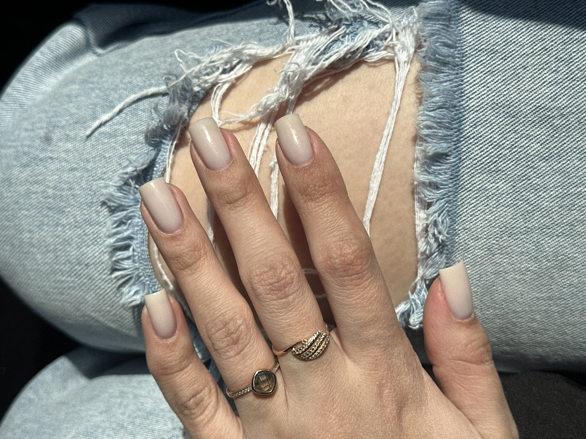 The image shows a close-up of a hand resting on a pair of distressed blue denim jeans. The jeans have rips and frayed edges, revealing the skin underneath. The hand features painted nails in a neutral beige color. Two rings adorn the fingers: one is a thin band with a round, dark-colored stone, and the other is a gold-colored band with a textured design. The background beyond the jeans is dark and indistinct. There is no evidence in the image to suggest the location in the city of Chaikovsky, Russia.