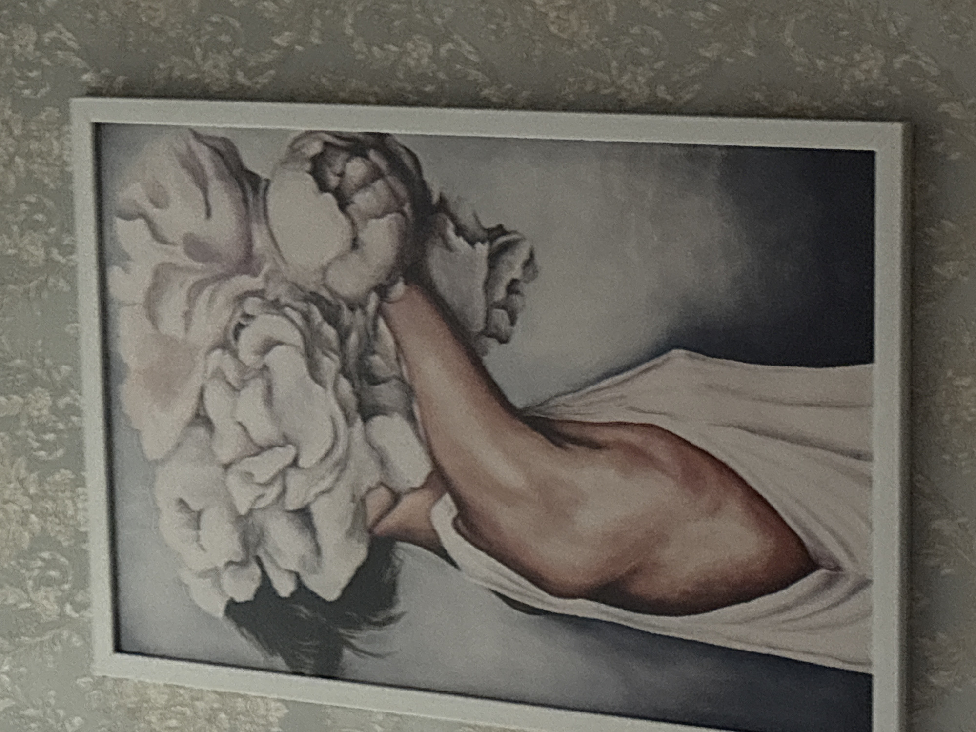 A framed painting is displayed on a textured wall in Rodniki, Russia. The painting depicts a woman whose head is obscured by a large arrangement of white flowers, possibly peonies. The woman is wearing a white tank top, and her bare shoulder and upper arm are visible. The background of the painting is a dark, muted gray. The wall behind the painting has a light-colored, floral-patterned wallpaper.
