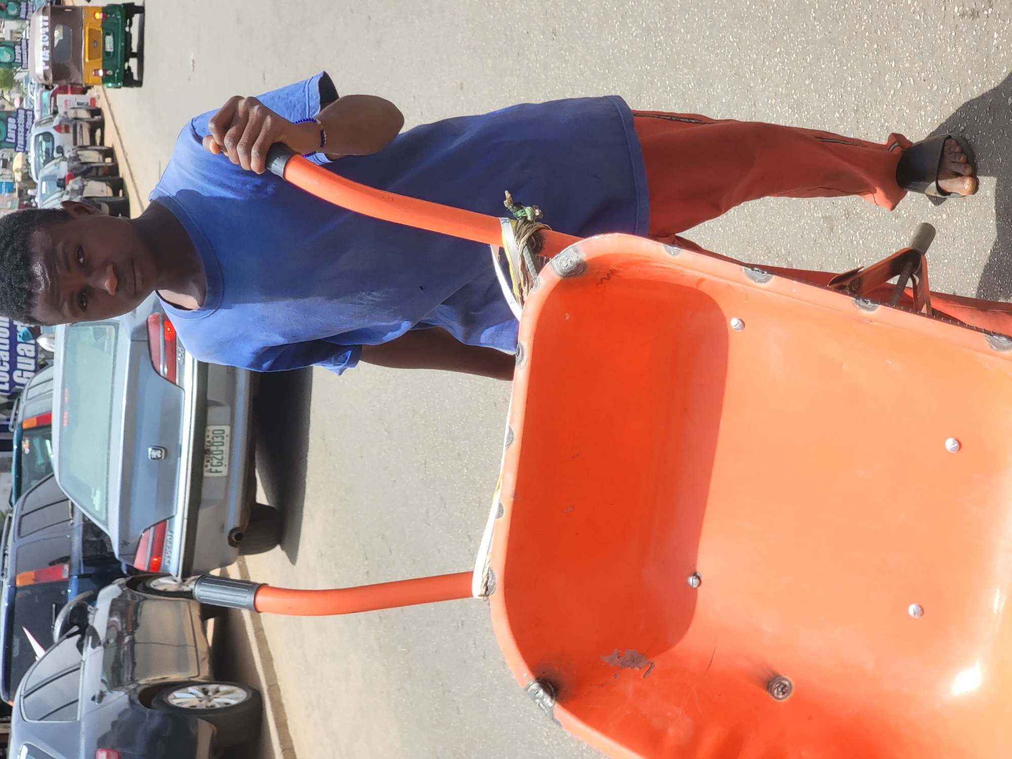 A young man in Jos, Nigeria, stands on an asphalt road, pulling an orange wheelbarrow towards the viewer. He wears a blue t-shirt, orange pants, and sandals. The wheelbarrow appears heavily used, with visible welds and repairs. Parked cars line the street behind him. Other vehicles, including a tricycle taxi, are visible further down the road. The license plate on the silver vehicle reads "FGZ0 030".