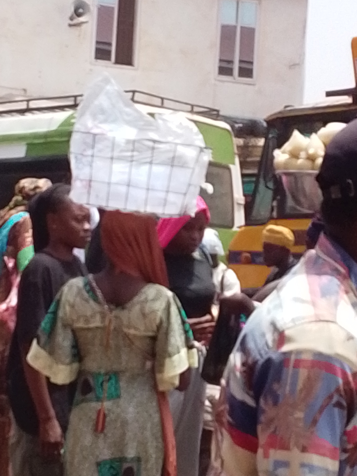 A crowded scene in Tamale, Ghana, is captured with a focus on individuals navigating a bustling environment.
The focal point is a woman carrying a large white object, possibly packaged goods, balanced on her head. She is wearing a patterned dress and a brown head covering. To her left stands another woman in a black shirt. Another woman with a pink head covering is slightly behind the woman carrying the load.
To the right, a man in a colorful patterned shirt stands with his back to the viewer. Behind these individuals are vehicles; a green van and a yellow vehicle carrying sacks of goods. A man in a yellow hat is inside the yellow vehicle.
The background reveals a building with two windows. The overall environment suggests a busy marketplace or street scene in the city of Tamale, Ghana.