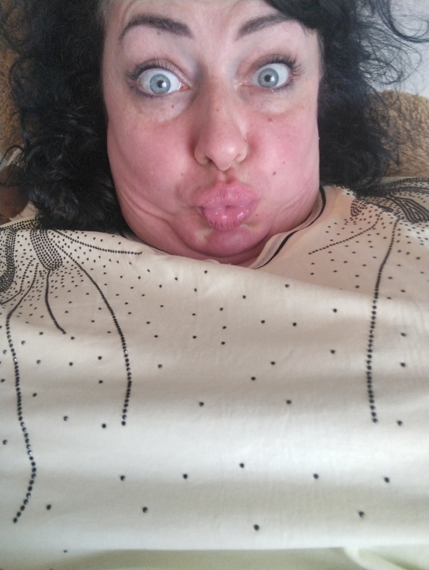 A woman with curly dark hair, light eyes, and a pouting expression is facing the camera. Her face is flushed, and she appears to be lying down, as the camera is positioned above her. She is wearing a light-colored garment adorned with black dotted patterns. There is no other information about the location or specific actions taking place.