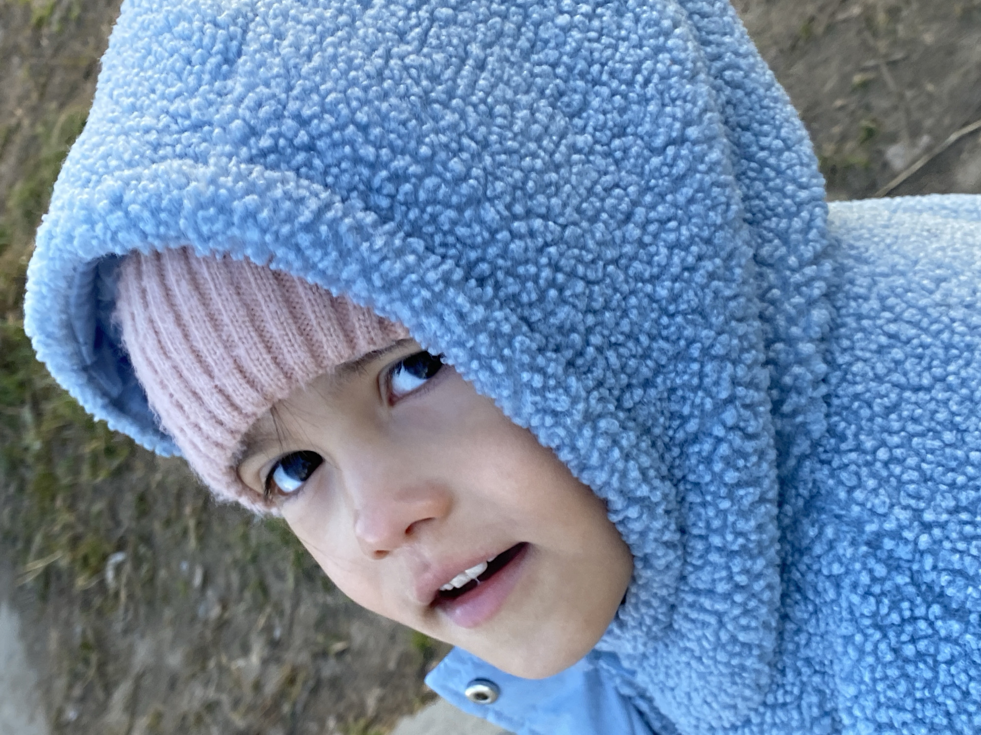 A close-up, high-angle shot depicts a young child wearing a light blue, fleece-like hooded jacket and a light pink knitted hat underneath. The child's face is partially framed by the hood and hat, with their eyes looking upwards and slightly to the left. The child has fair skin, dark eyes, and a slightly open mouth, revealing a few teeth. The background is slightly blurred, showing hints of a natural, possibly outdoor environment with muted colors. The clothing suggests a cool weather setting. There is no information to indicate that this image was taken in the city of Chaikovsky, Russia.
