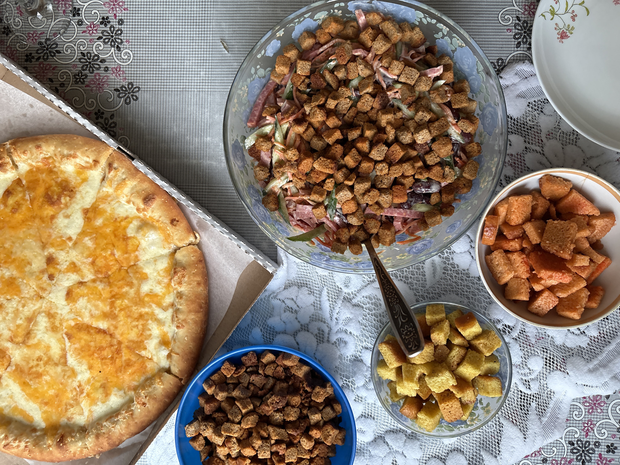 A top-down shot shows a table laden with food, possibly in Ust-Ilimsk, Russia. A pizza in a cardboard box sits on the left. To the right of the pizza is a salad in a large glass bowl, topped with croutons and containing sliced meat and vegetables. A serving spoon rests in the bowl. Three smaller bowls are arranged around the large salad bowl. One bowl is blue, one is glass, and one is white with a colorful rim. Each smaller bowl contains croutons of different colors. To the right of the salad bowl are a stack of white plates. The table is covered with two tablecloths, one with a floral pattern and one with a white lace pattern.