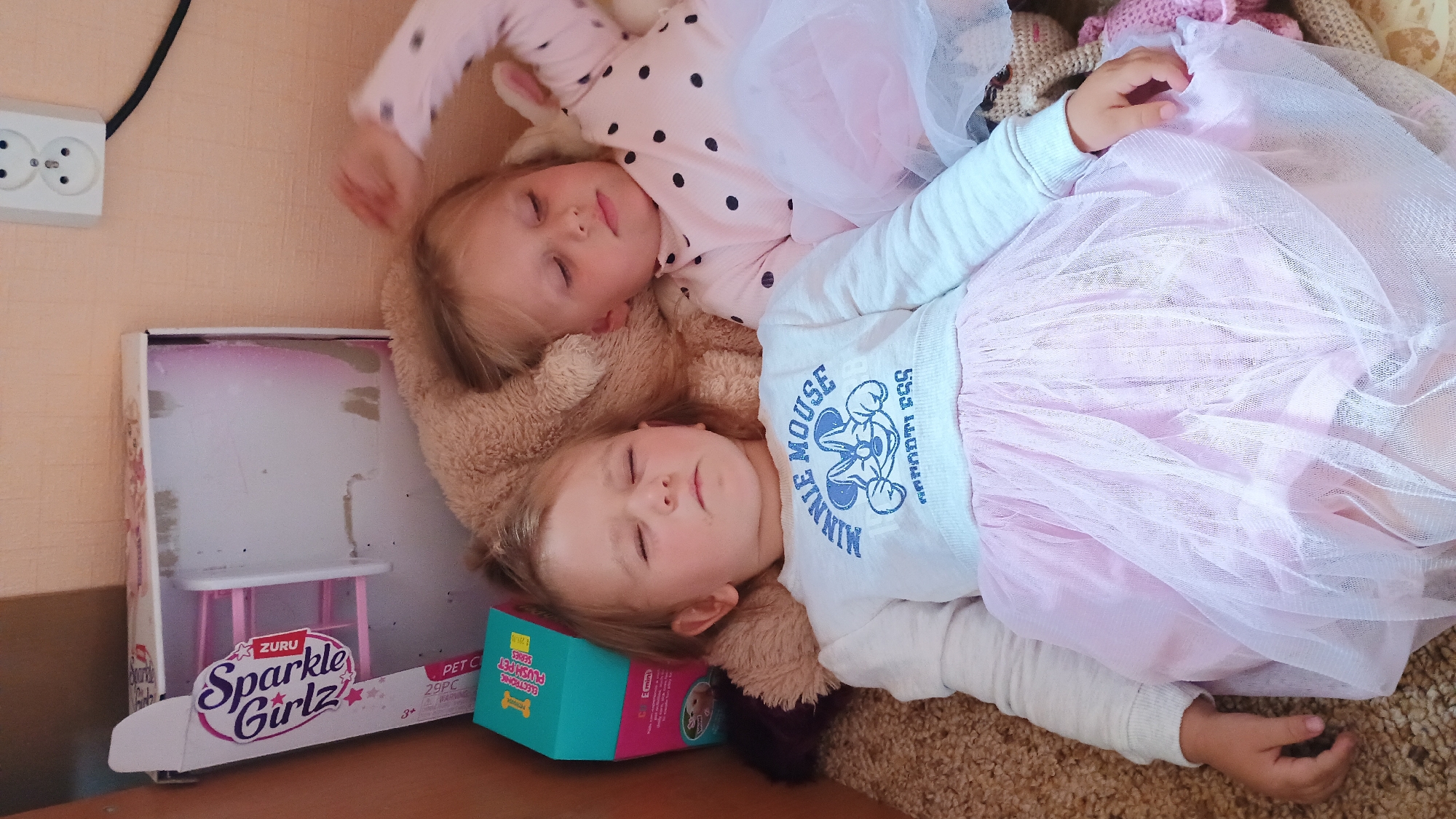 Two young girls are sleeping side-by-side on a fuzzy, beige surface, possibly a carpet or blanket. The girl on the left, wearing a white sweatshirt with a blue "Minnie Mouse" graphic and a pink tutu, has her eyes closed. The girl on the right, wearing a pink polka-dot shirt, also has her eyes closed. Behind the girls, a large, light brown stuffed toy, possibly a bear, is visible. To the left of the girls, a partially open cardboard box with "Sparkle Girlz" branding is present, along with a small teal box. A white electrical outlet with two sockets is mounted on a peach-colored wall in the upper right corner of the image, with a black cord extending from it. The scene appears to be indoors, in what could be a living room or bedroom. The location is not specified in the image or its context.
