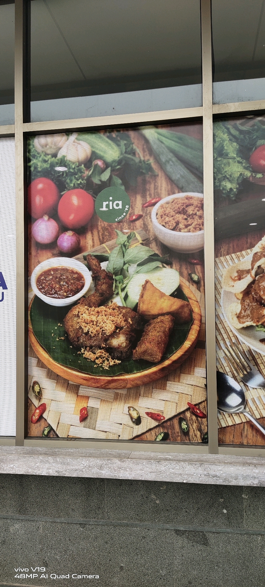 The image displays a framed advertisement, likely a window decal, for "ria since '98," a restaurant or food service in Pekanbaru, Indonesia. The advertisement features a close-up, overhead view of a dish of fried chicken served with rice, chili sauce, and various vegetables, including tomatoes, onions, and cucumber. The food is arranged on a banana leaf within a woven wooden bowl, set against a backdrop of wooden planks. Several red and green chili peppers are scattered around the dish. The advertisement is partially visible, with the left side obscured by a white textured surface and the right side by another similar advertisement. The lower portion of the image shows a concrete ledge and a rough concrete wall, consistent with an outdoor urban environment in Pekanbaru. The "vivo V19 48MP AI Quad Camera" watermark indicates the image was captured with this device.