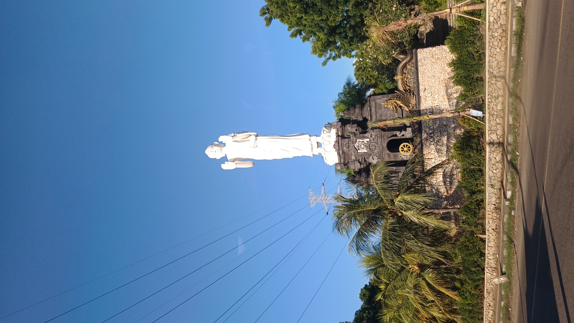 The image shows a standing white Buddha statue in Melaya, Indonesia. The statue is positioned atop a decorative structure with Balinese elements and a stone facade. Palm trees and other vegetation surround the structure. Power lines intersect the clear blue sky behind the statue. A road runs along the right edge of the image.