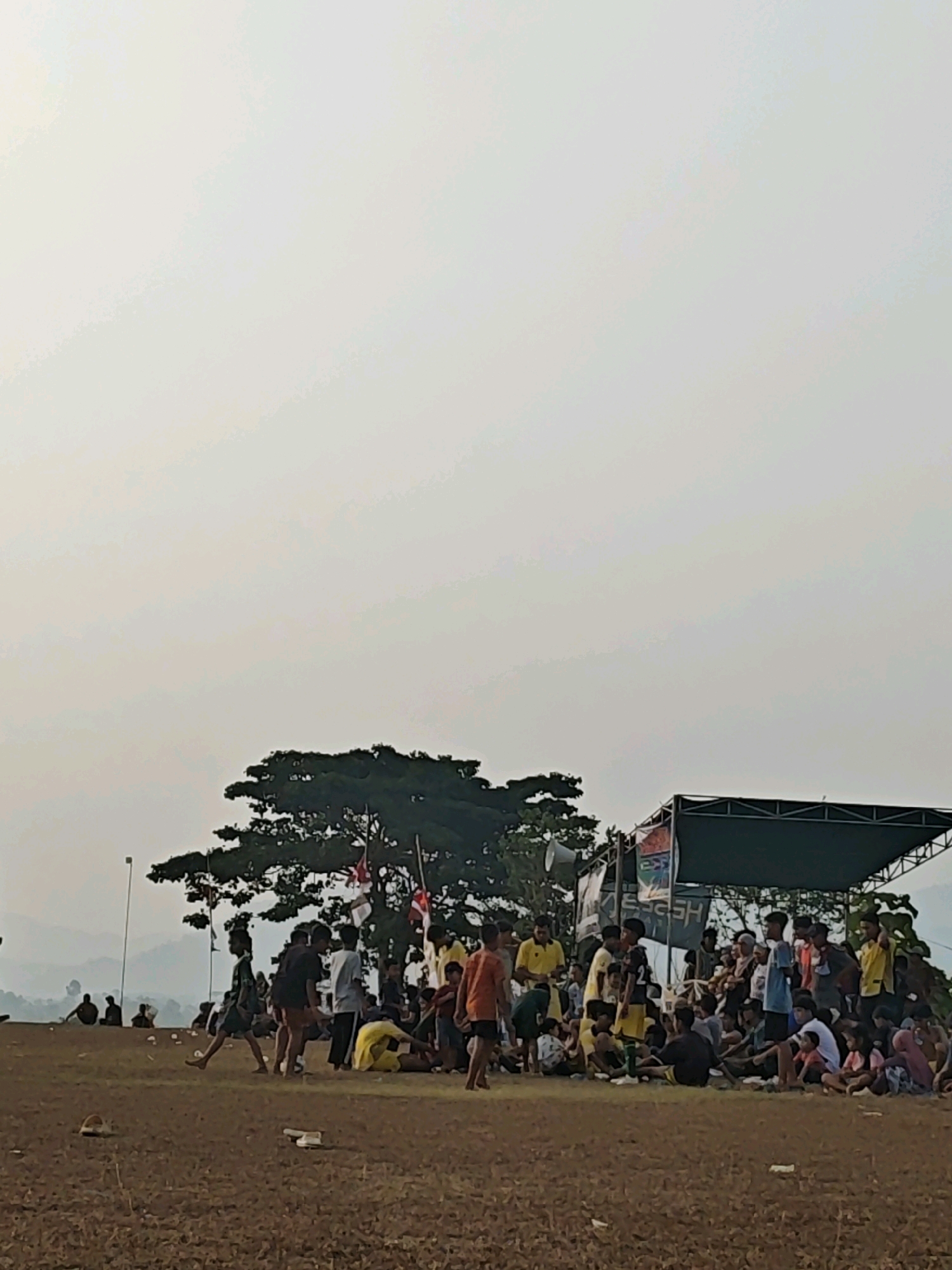 The image depicts an outdoor scene in Karang Sari, Indonesia, likely a community event or gathering. In the foreground, the ground is covered in dry, brown dirt with scattered litter. A group of people, predominantly young males, are gathered in the middle ground. Some are standing, some are sitting on the ground, and a few are in motion. They are dressed in casual clothing, with some wearing athletic attire.
Behind the crowd, a large tree with dark green foliage stands prominently, its branches reaching towards the sky. To the right of the tree, a stage or platform is set up, covered with a dark green canopy. Banners are visible on the stage, though the text is largely illegible. Several loudspeakers are mounted on poles near the stage. In the background, rolling hills are faintly visible through a hazy, light-colored sky, suggesting a rural or semi-rural setting. The overall atmosphere appears to be one of casual congregation.