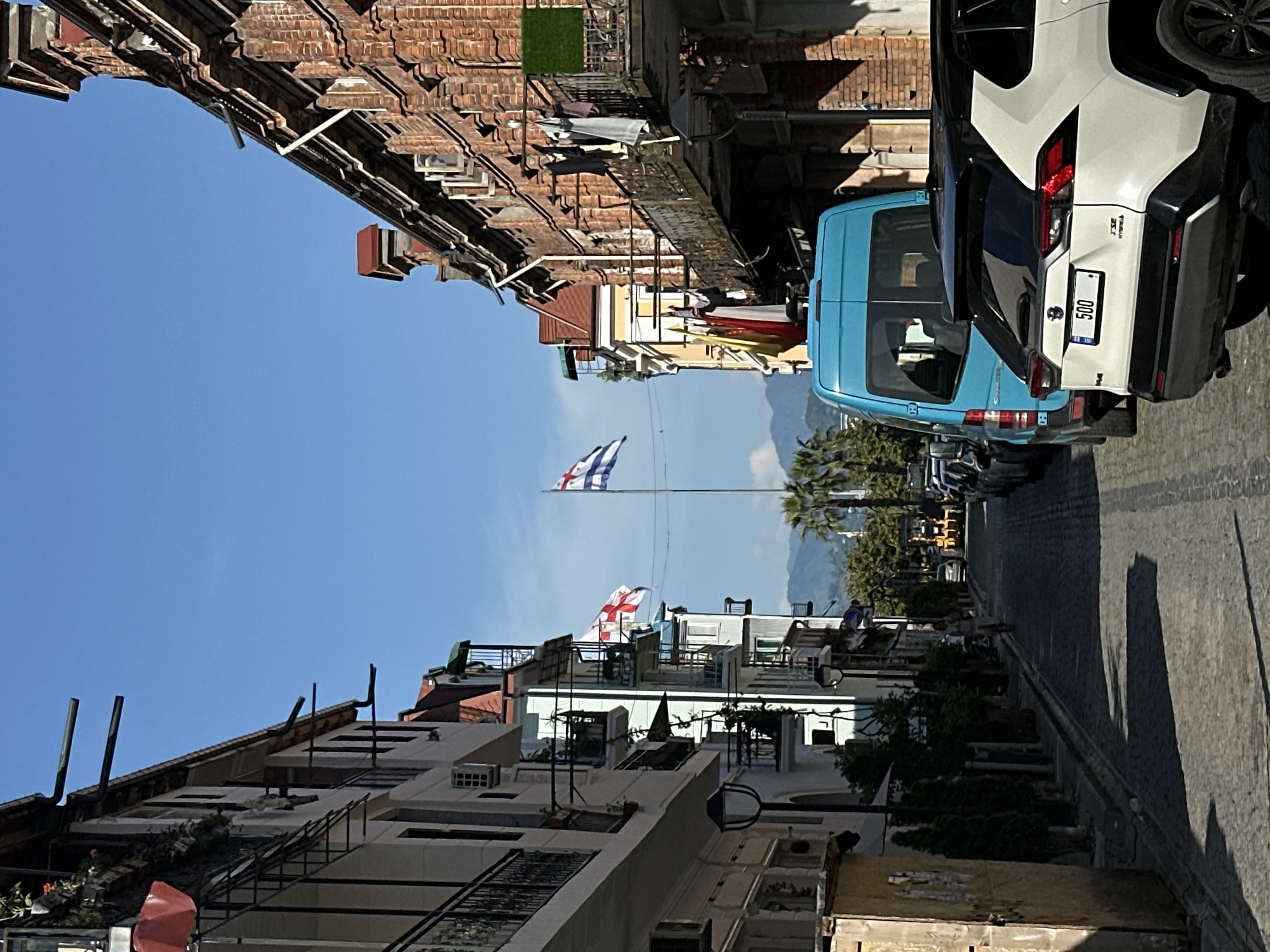 The photograph shows a street scene in Batumi, Georgia. Buildings of varying heights with mixed architectural styles line the narrow street. A Georgian flag flies from a tall pole in the distance, while another flag flies from a building closer to the viewer. A light blue van and a white SUV are parked along the right side of the street. Several other vehicles can be seen further down the road. Palm trees and distant mountains are visible beyond the buildings. The sky is clear and blue. Laundry hangs from lines strung between some of the buildings.