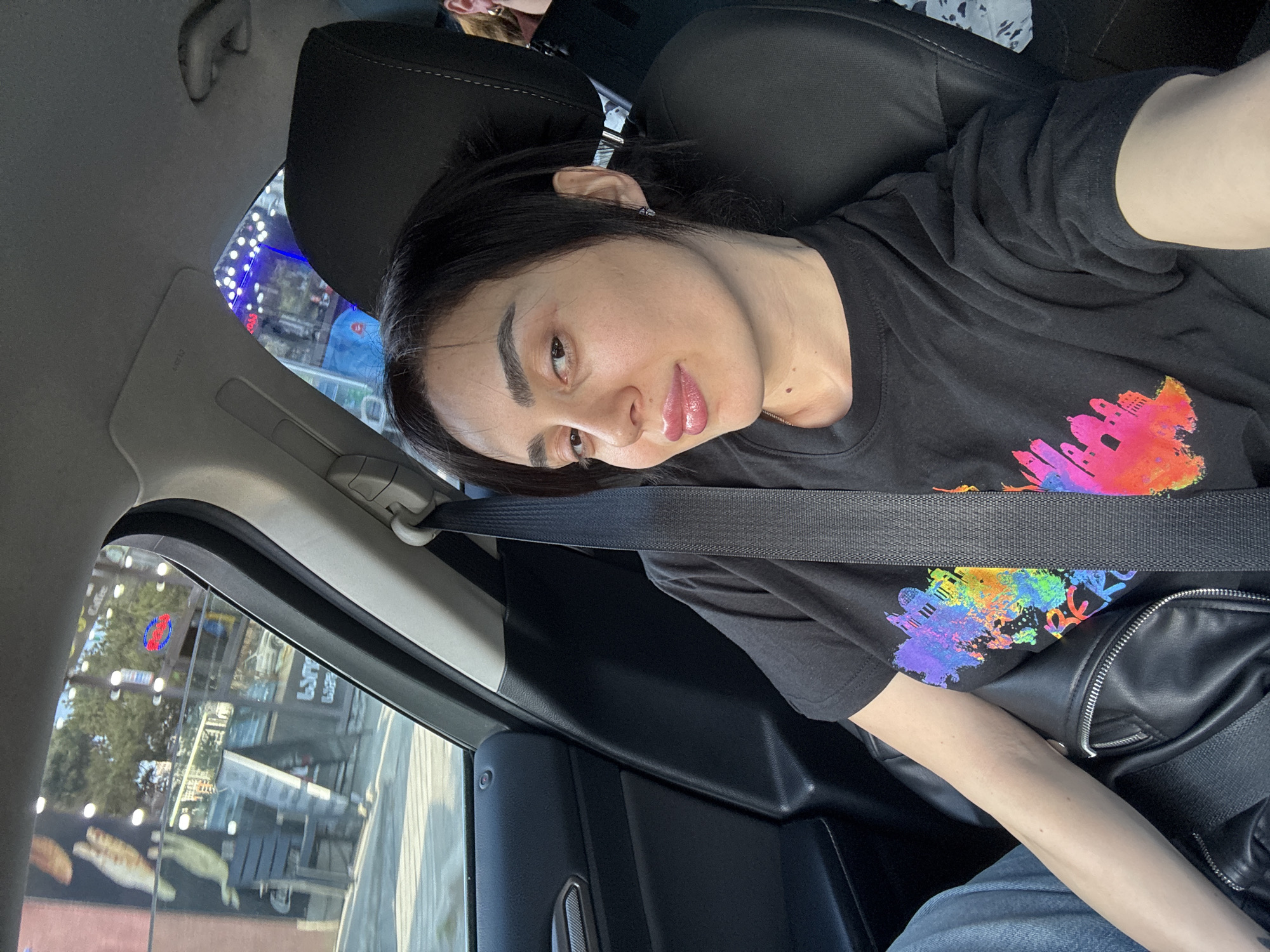 The image shows a person in the passenger seat of a car in Tbilisi, Georgia. The person has dark hair and is wearing a black t-shirt with a colorful graphic. A seatbelt is across their chest. Outside the car window, the architecture of Tbilisi can be seen, including businesses and street lights.